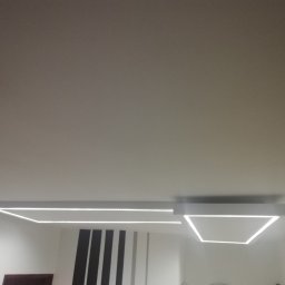 Look home remonty i instalacje - Nowoczesne oświetlenie sufitowe z białymi, geometrycznymi oprawami LED, widoczne na tle białego sufitu i ściany z dekoracyjnymi pionowymi pasami.