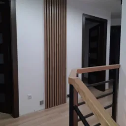 Nowoczesny przedpokój z drewnianą ażurową ścianką działową i schodami z drewnianą balustradą, widoczne drzwi wewnętrzne z ciemnego drewna.