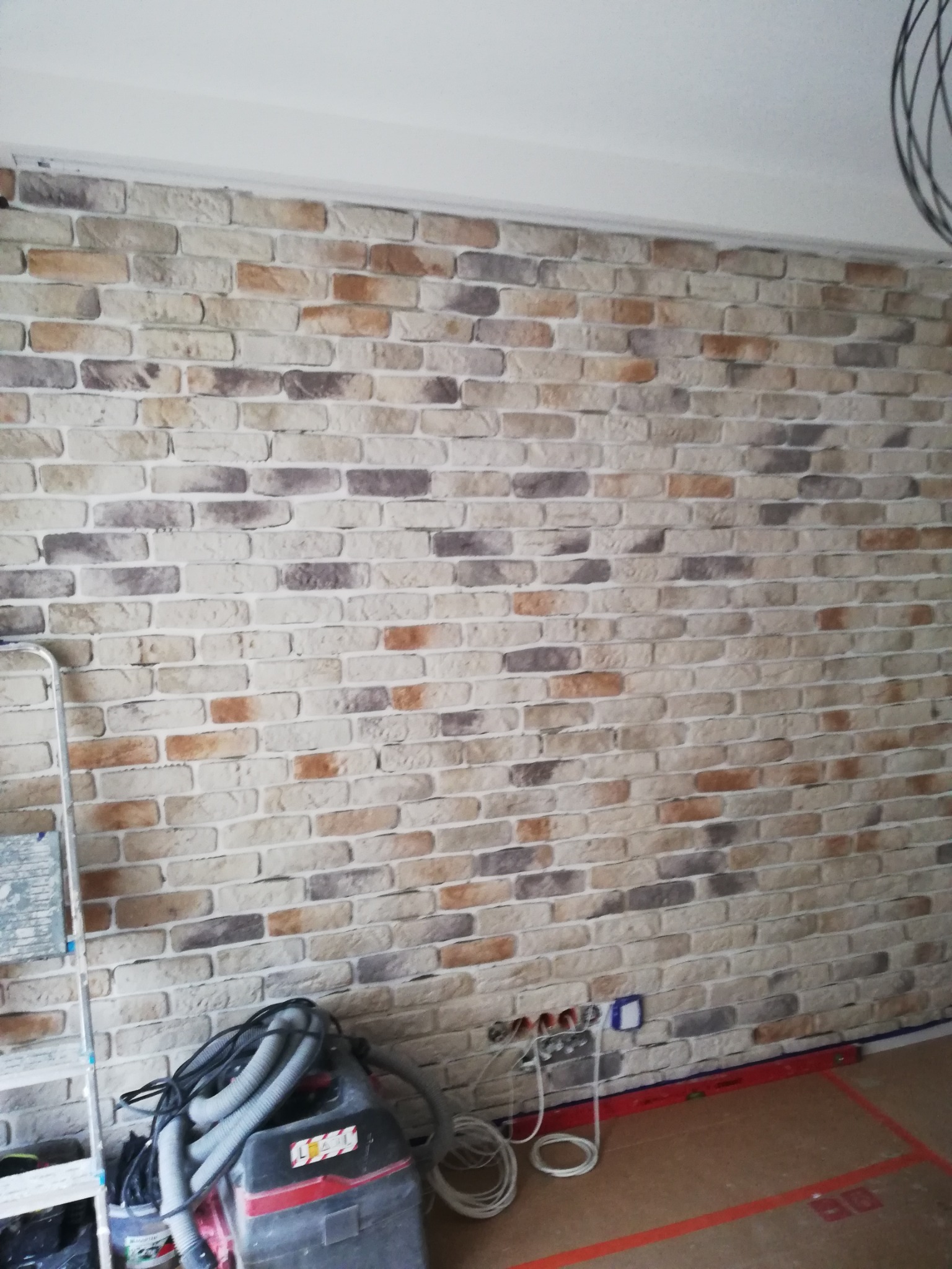 Ściana wykończona dekoracyjną cegłą w odcieniach beżu, brązu i szarości, widoczne gniazdka elektryczne, poziomica, odkurzacz przemysłowy i drabina, sugerujące prace wykończeniowe w toku.