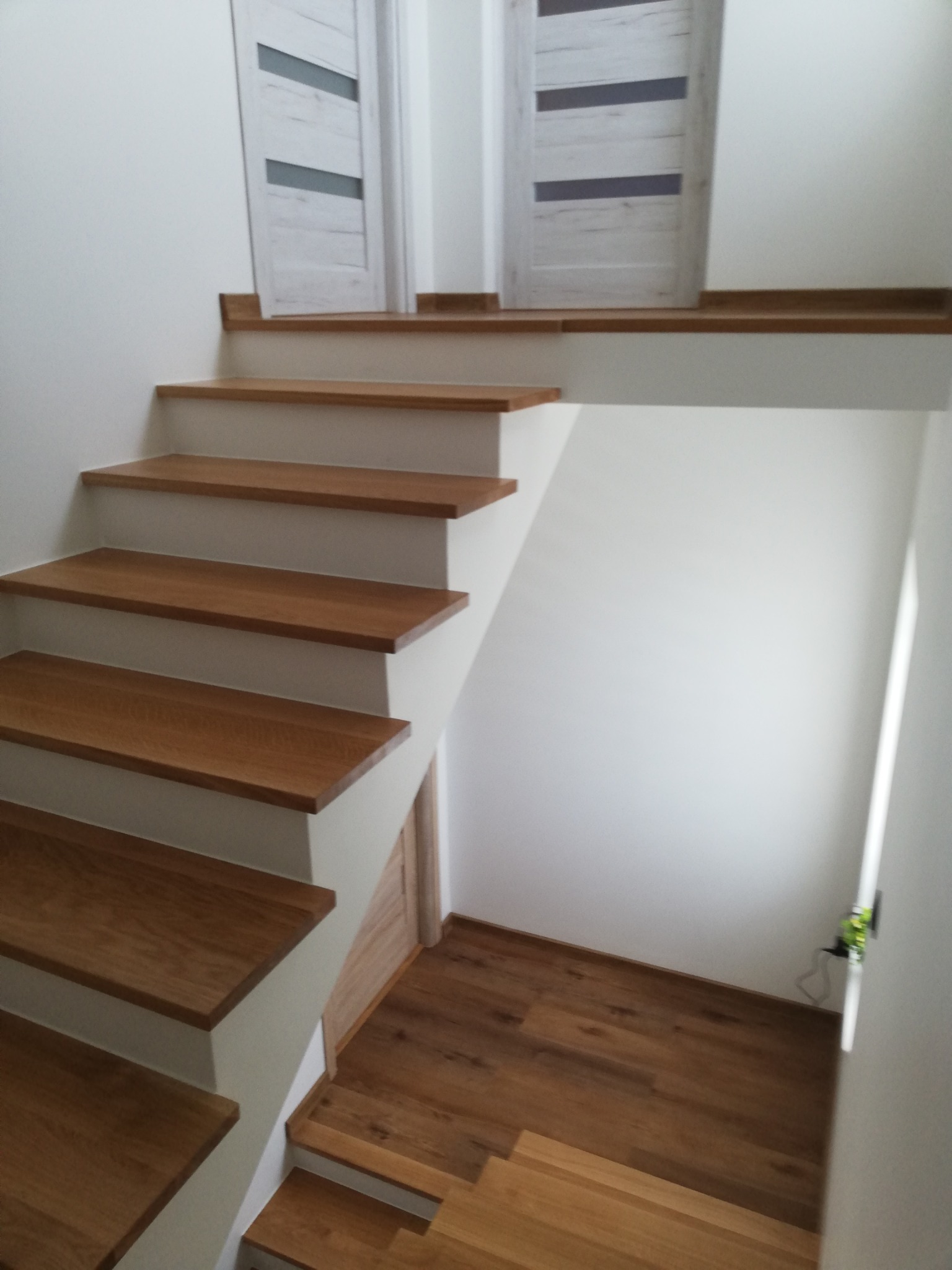 Drewniane schody z białymi podstopnicami prowadzące do dwóch białych drzwi z szarymi wstawkami, widok z dołu.