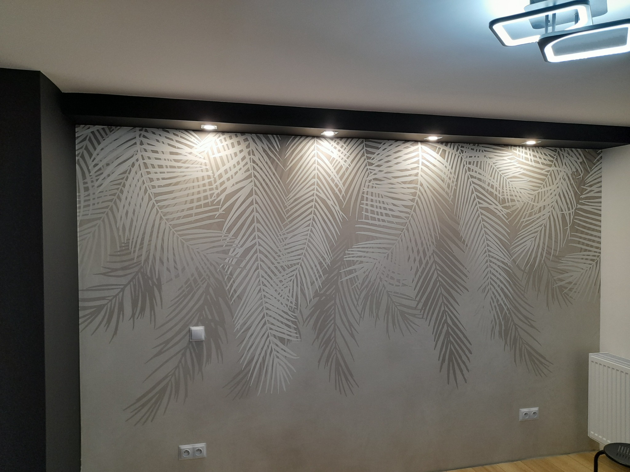 Ściana w pokoju z beżową tapetą w motywy liści palmowych, oświetlona halogenami w czarnej listwie podsufitowej i nowoczesną lampą, widoczne gniazdka elektryczne i włącznik światła.
