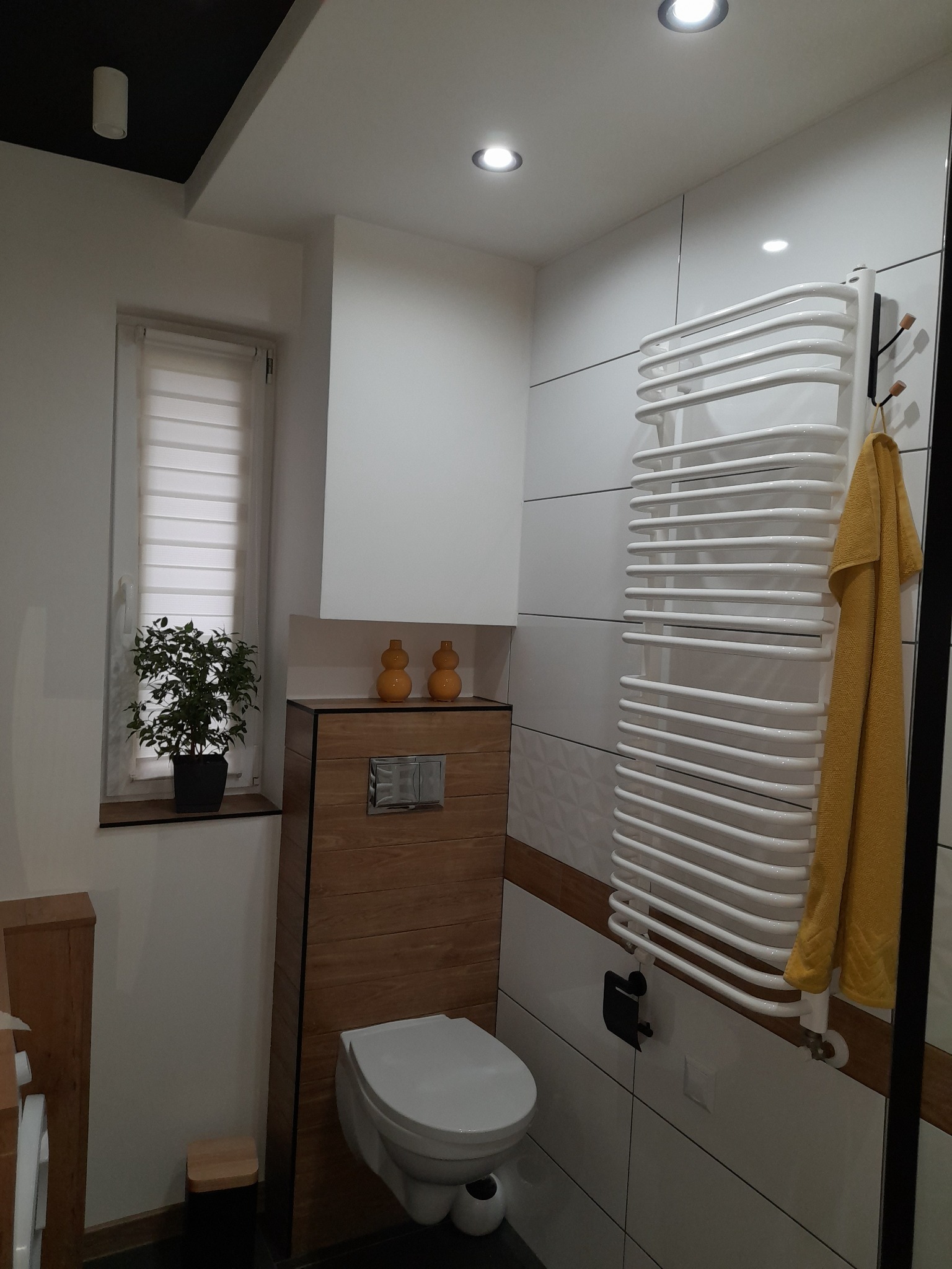 Nowoczesna łazienka z białymi płytkami, drewnianymi akcentami, grzejnikiem drabinkowym z żółtym ręcznikiem i rośliną doniczkową na parapecie.