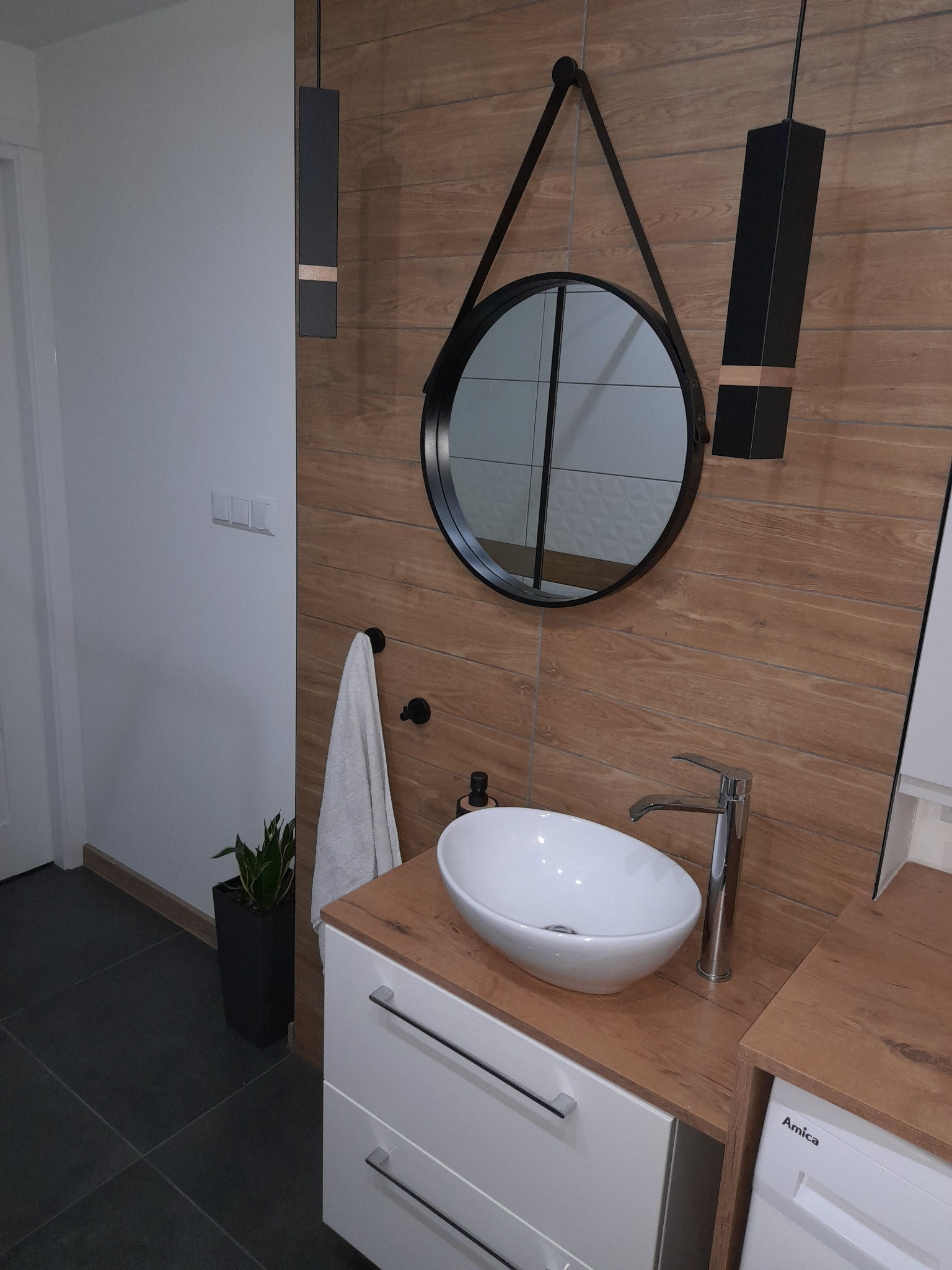 Nowoczesna łazienka z okrągłym lustrem na skórzanym pasku, umywalką nablatową i drewnopodobnymi płytkami ściennymi, uzupełniona minimalistycznymi lampami wiszącymi i rośliną w czarnej doniczce.