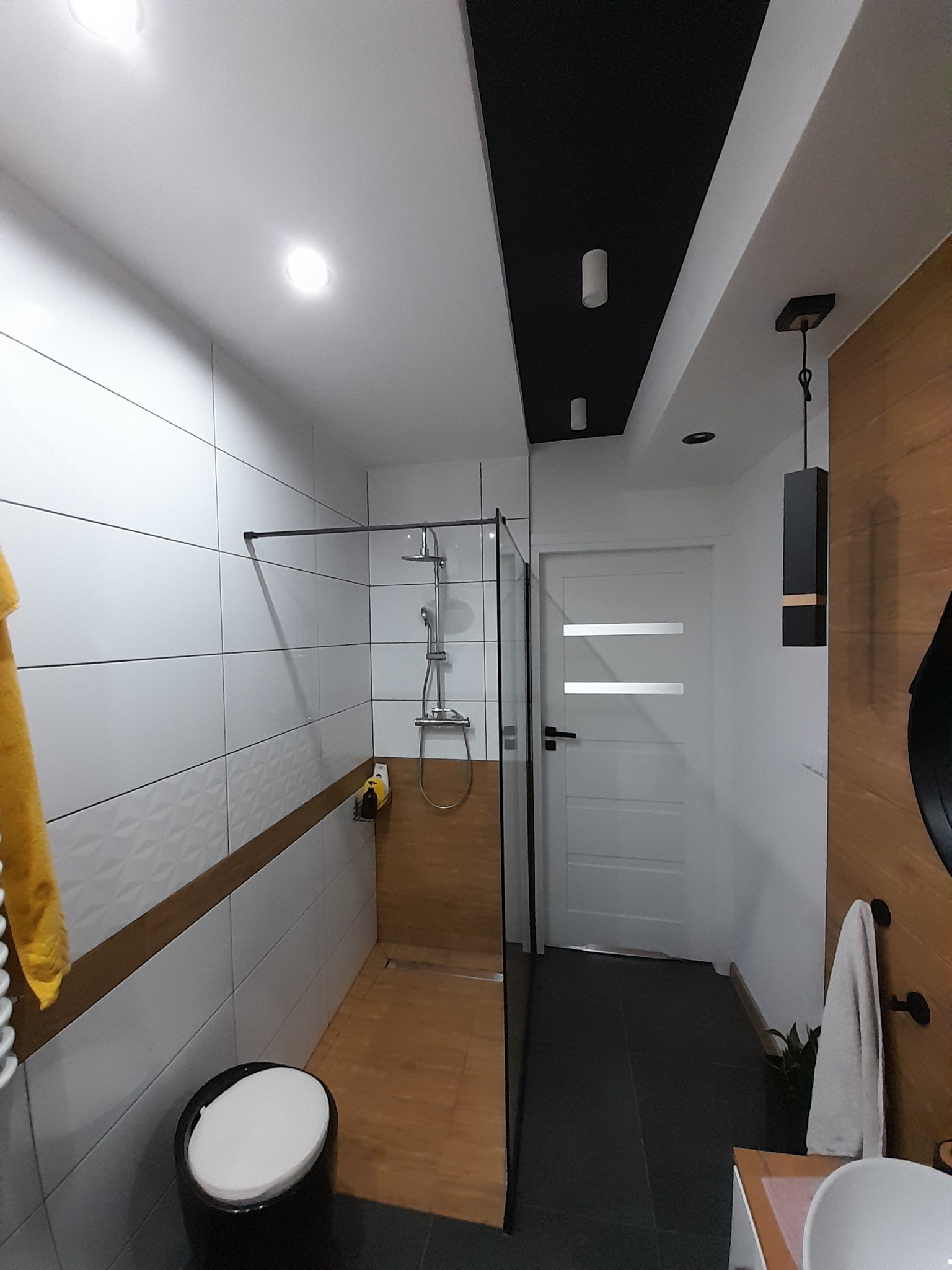 Nowoczesna łazienka z kabiną prysznicową typu walk-in, wykończona białymi płytkami z geometrycznym wzorem i drewnopodobnymi akcentami, czarną podłogą i sufitem, oświetlona punktowymi lampami...