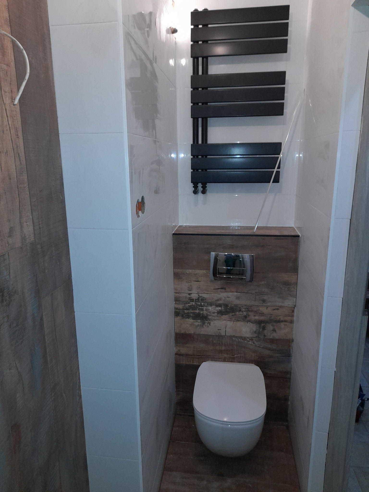 Nowoczesna toaleta w małej łazience z białymi płytkami imitującymi drewno, czarnym grzejnikiem drabinkowym i ukrytą spłuczką za panelem z drewnopodobnych desek.