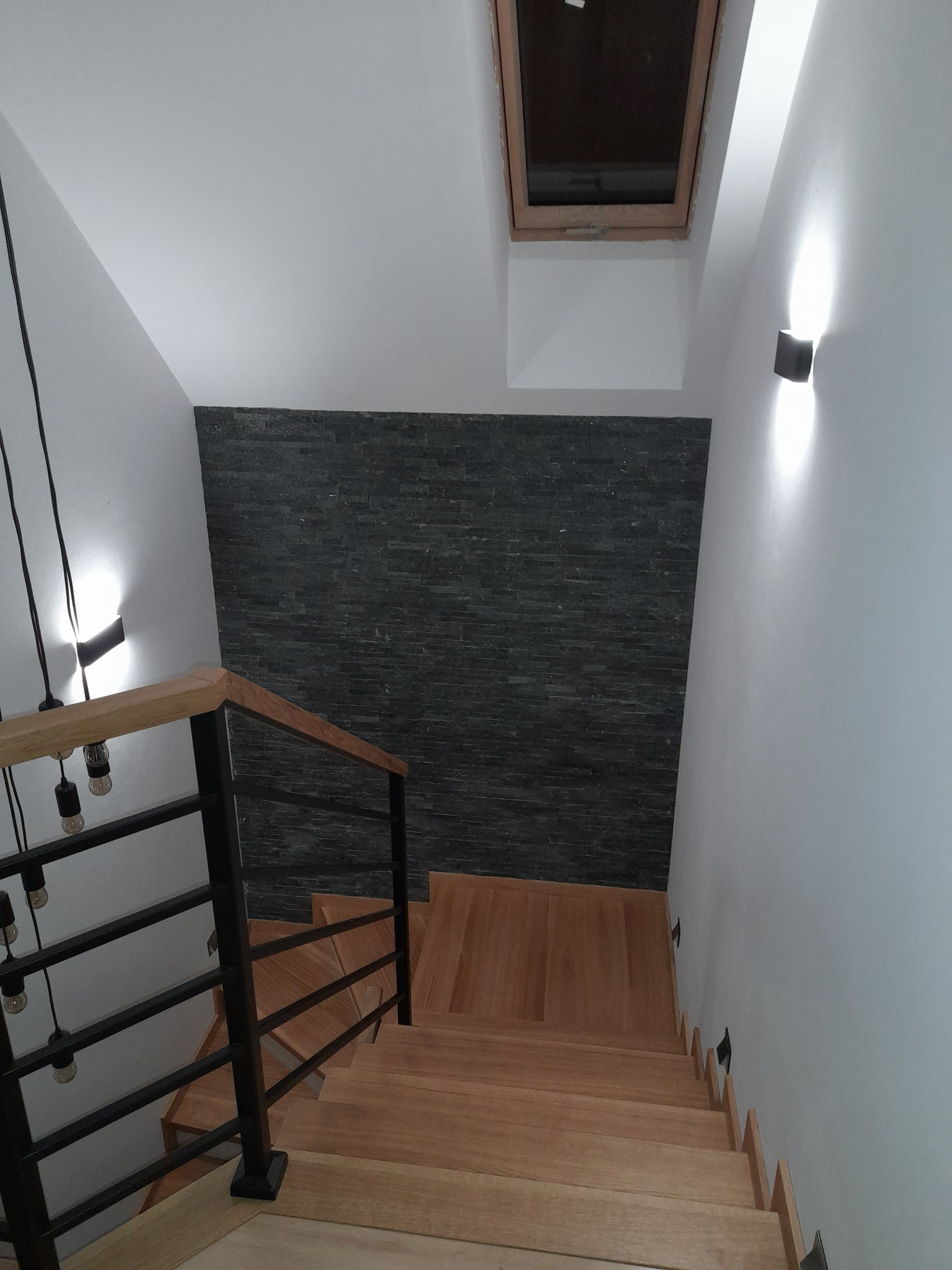 Nowoczesne drewniane schody z czarną balustradą i oświetleniem ściennym, ściana wykończona czarnym kamieniem dekoracyjnym, okno dachowe.