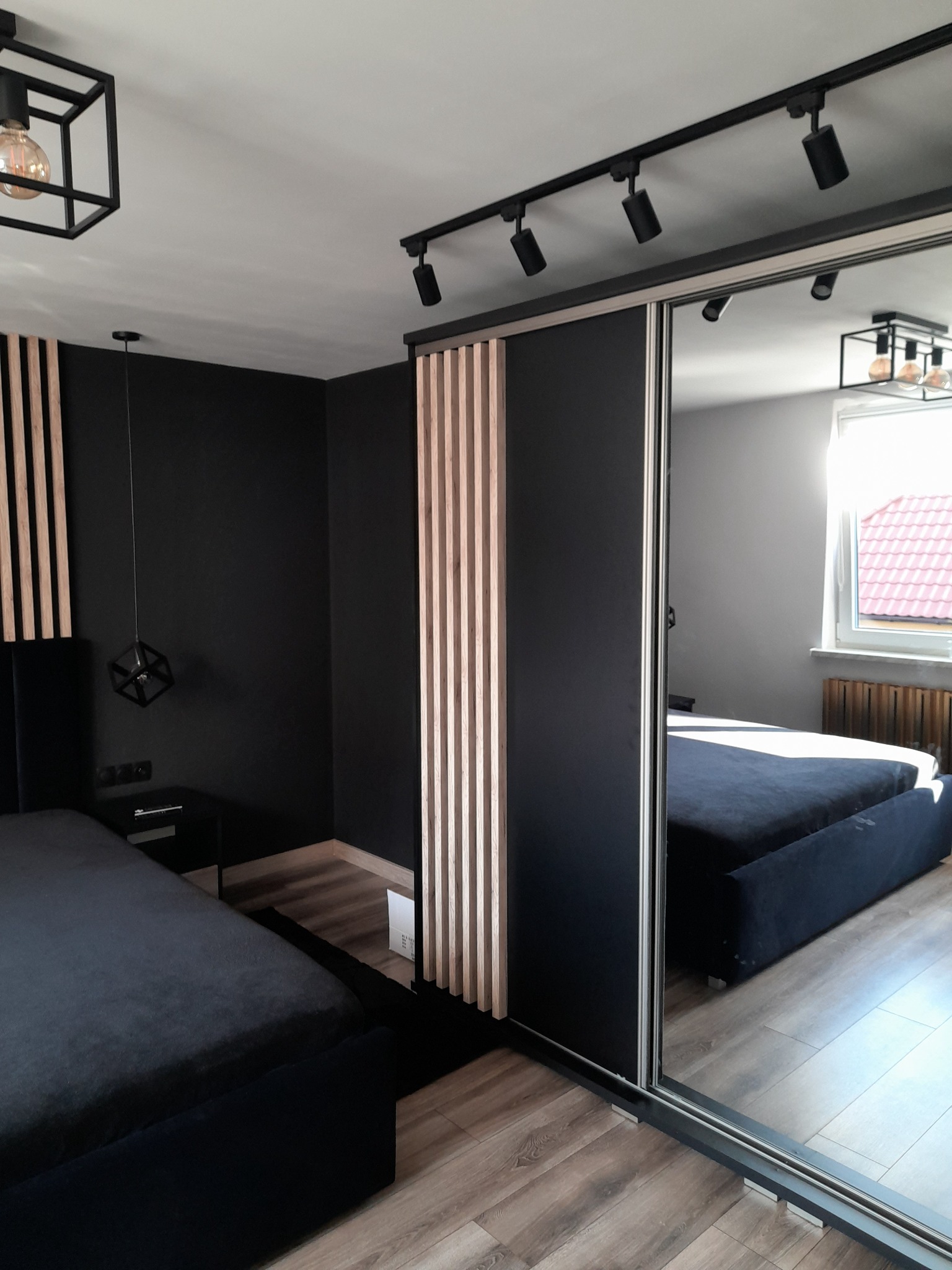 Nowoczesna sypialnia z czarnymi ścianami, drewnianymi lamelami, łóżkiem z ciemną tapicerką i szafą z lustrzanymi drzwiami przesuwnymi, oświetlona reflektorami szynowymi i lampą wiszącą...