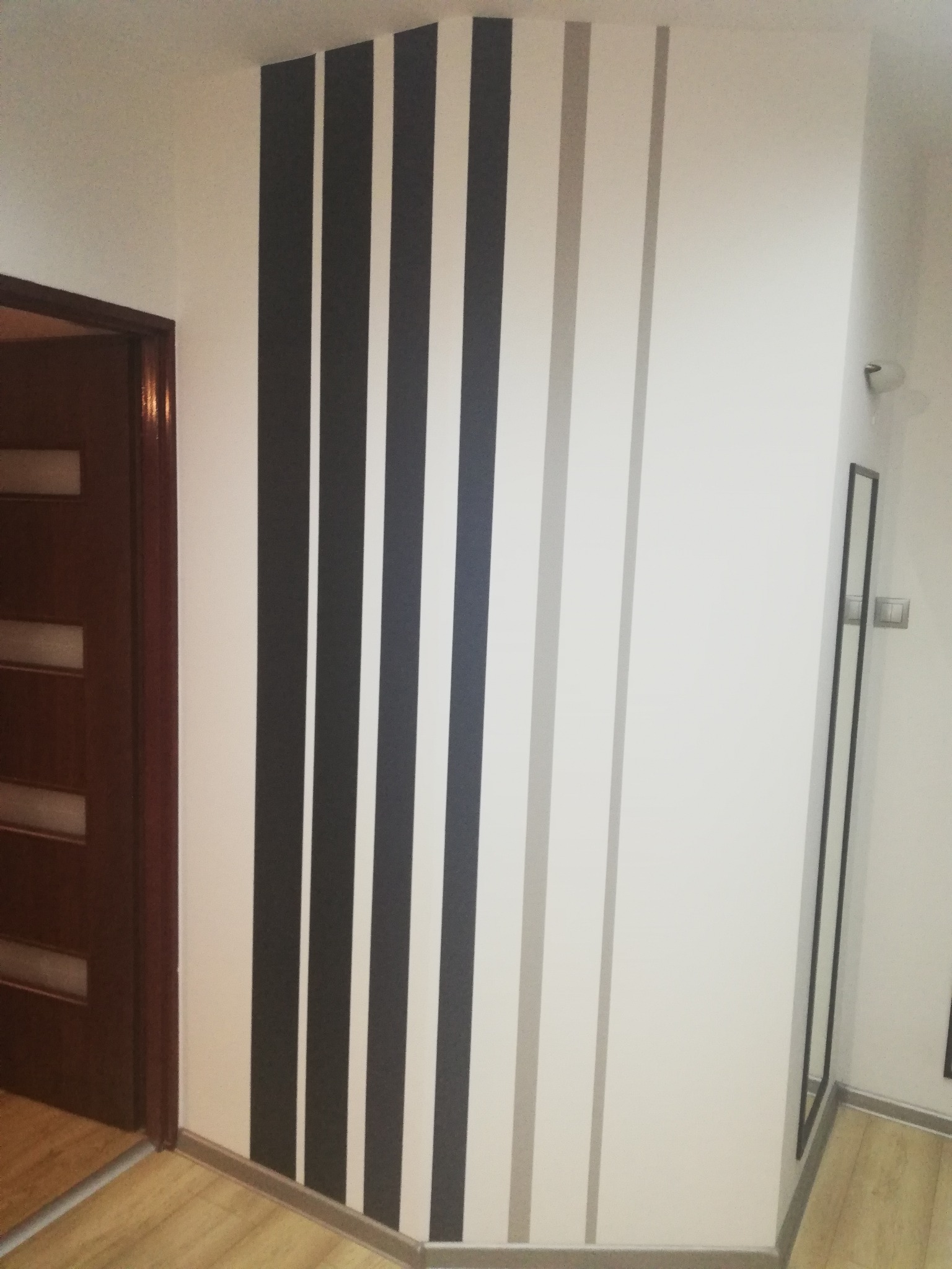 Pomysłowe malowanie ścian w pionowe pasy: czarne, białe i szare, w rogu pokoju widoczna drewniana rama drzwi i wysoka, wąska rama lustra.