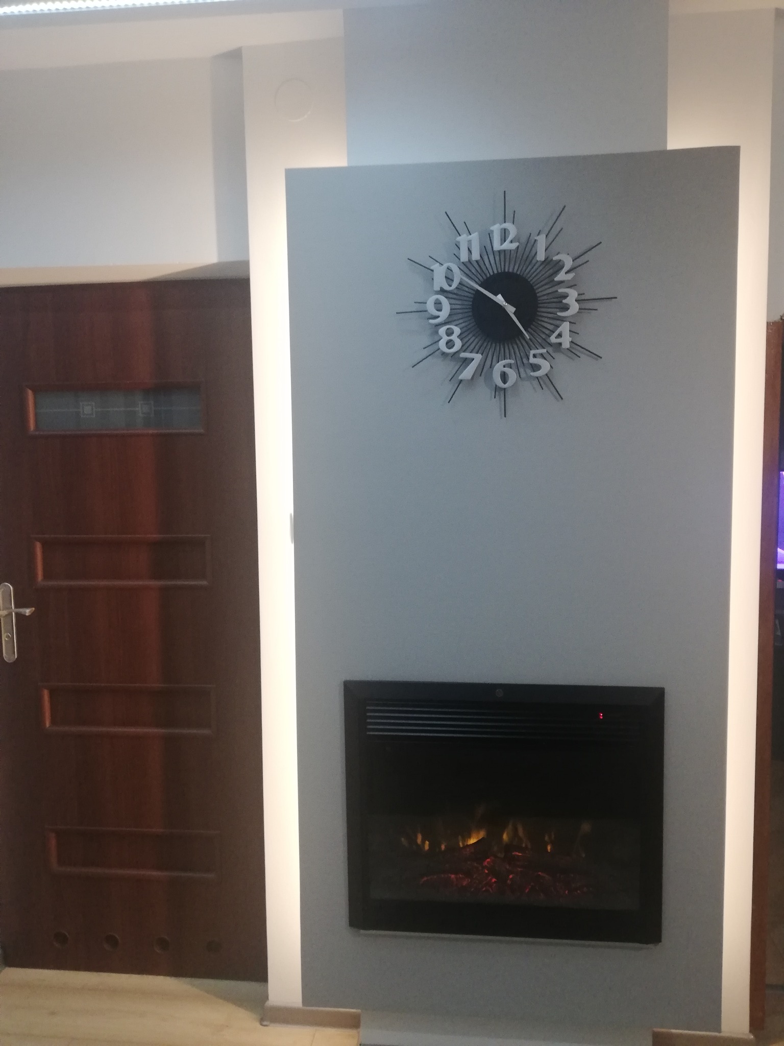 Szara ściana z elektrycznym kominkiem i zegarem słonecznym, obok drewniane drzwi z przeszkleniami, oświetlenie punktowe.