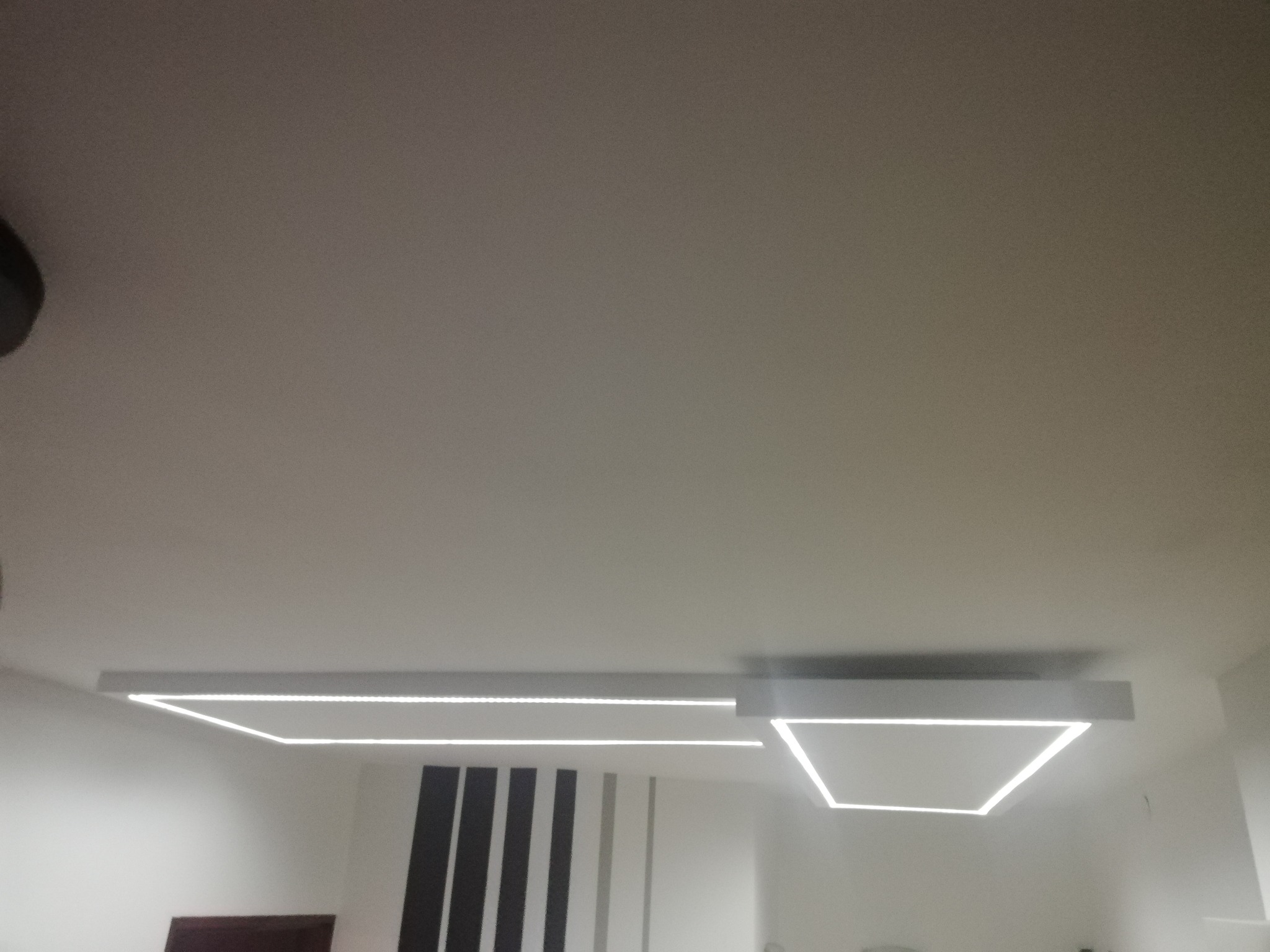 Nowoczesne oświetlenie sufitowe z białymi, geometrycznymi oprawami LED, widoczne na tle białego sufitu i ściany z dekoracyjnymi pionowymi pasami.