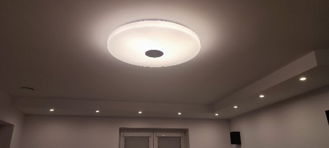 Biały sufit z okrągłą lampą LED z czarnym środkiem, dodatkowo oświetlenie punktowe w listwie na suficie podwieszanym.