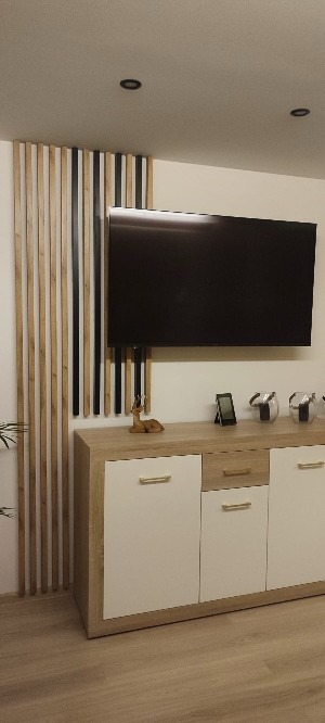Nowoczesny salon z drewnianymi lamelami na ścianie, telewizorem LCD, komodą z białymi frontami i złotymi uchwytami, oraz dekoracjami na komodzie.