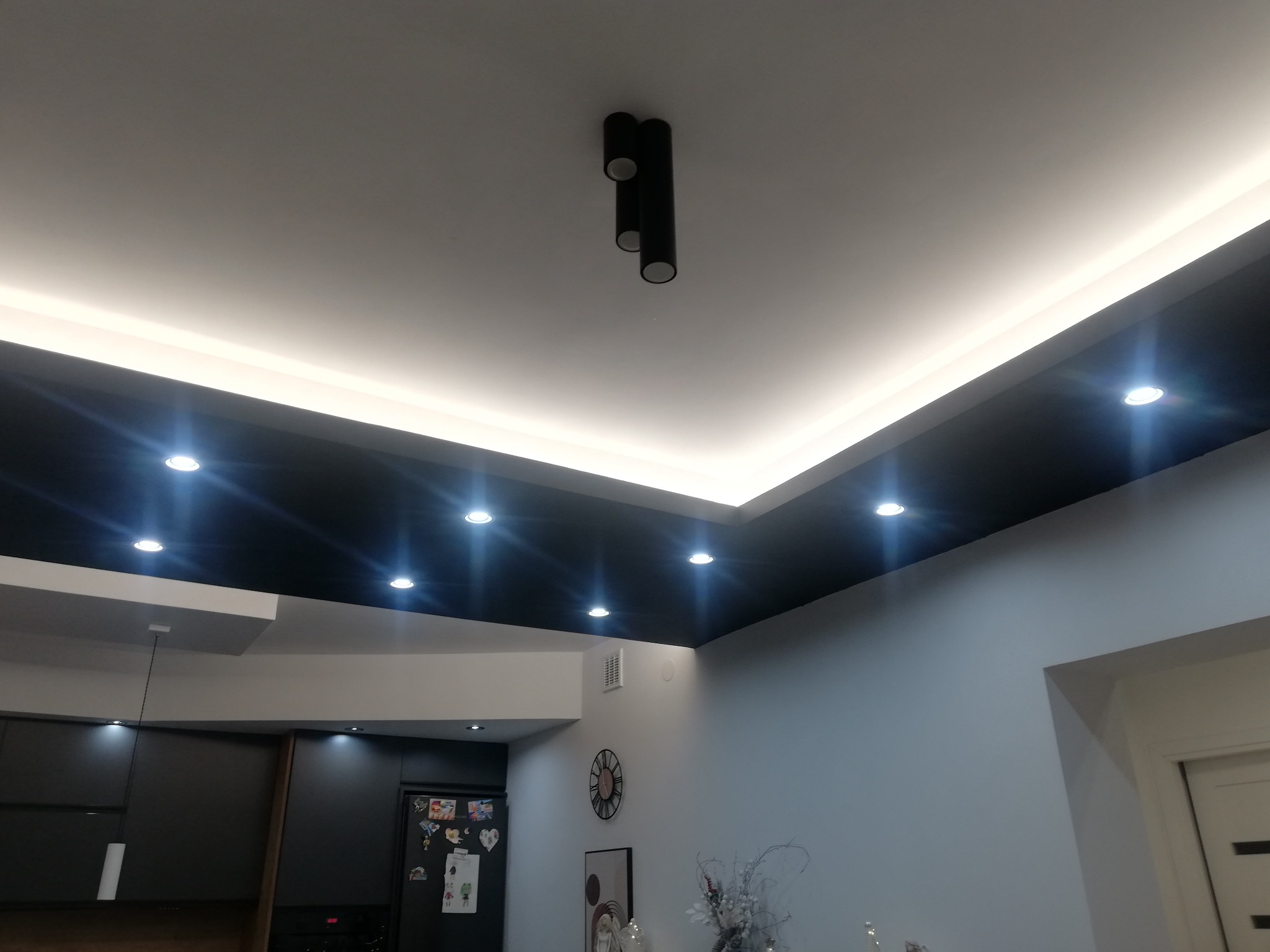 Nowoczesne oświetlenie sufitowe z czarnym podwieszanym sufitem i białymi panelami LED, widok z dołu na minimalistyczną lampę tubową i fragment zabudowy kuchennej w tle.