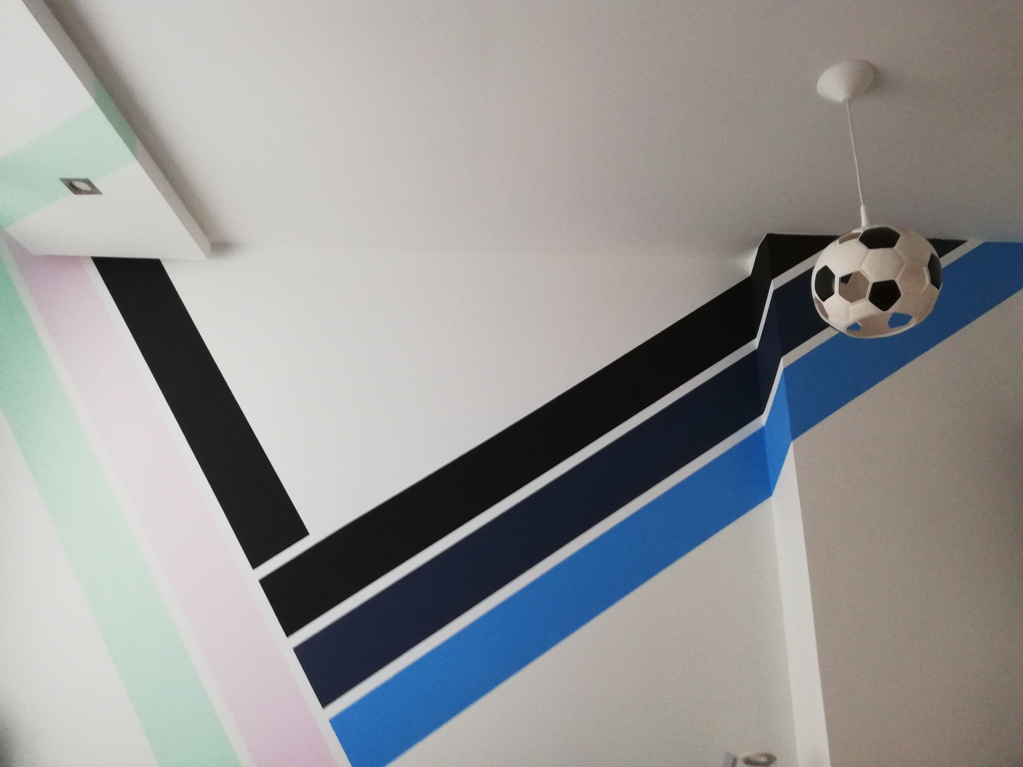Pokój dziecięcy z geometrycznym malowaniem ścian w odcieniach pastelowych i niebieskich, uzupełniony lampą sufitową w kształcie piłki nożnej.