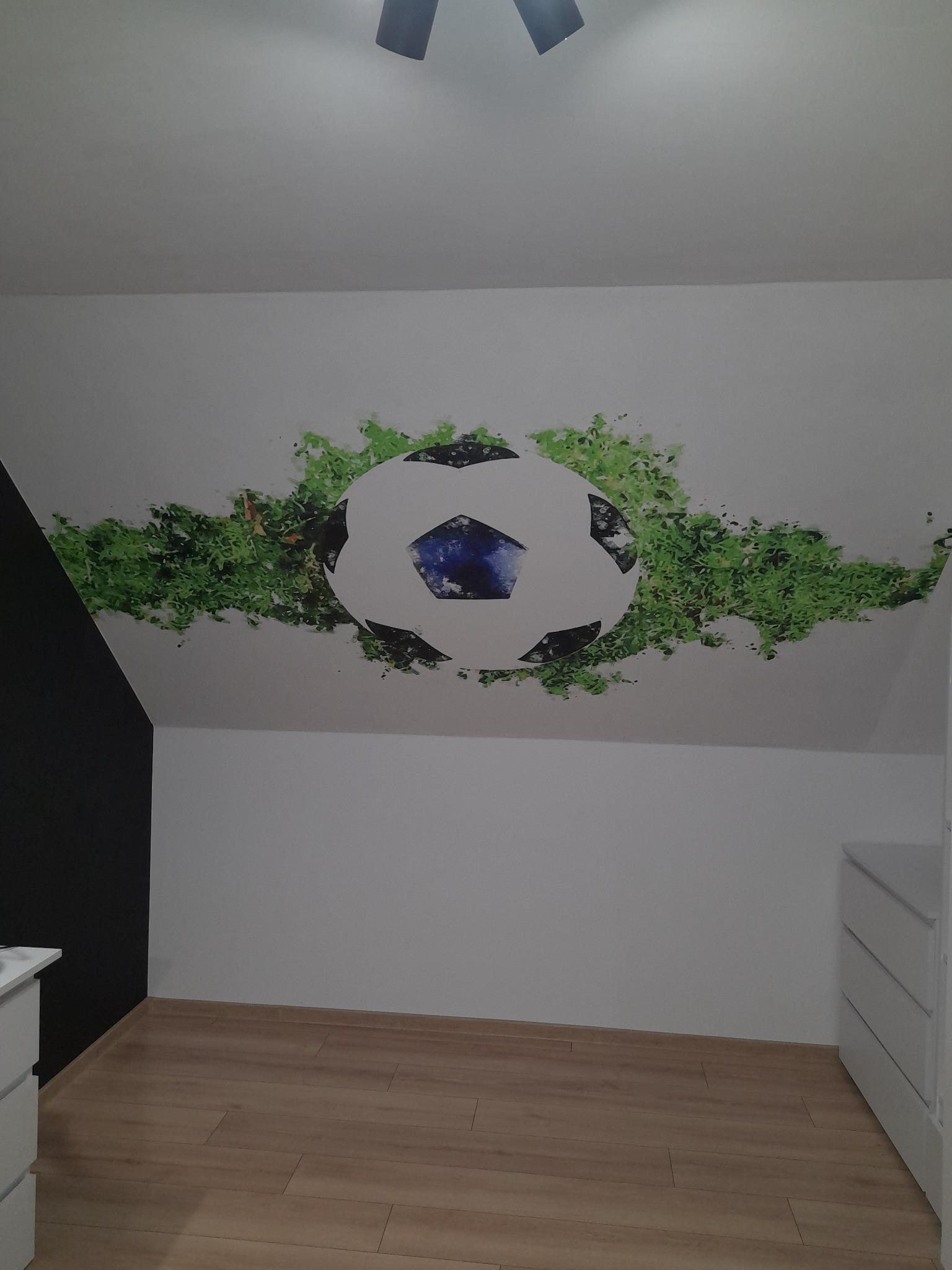 Pokój dziecięcy na poddaszu z drewnianą podłogą i białą komodą; na skosie sufitu widoczny malunek piłki nożnej otoczonej zielonymi elementami imitującymi trawę.