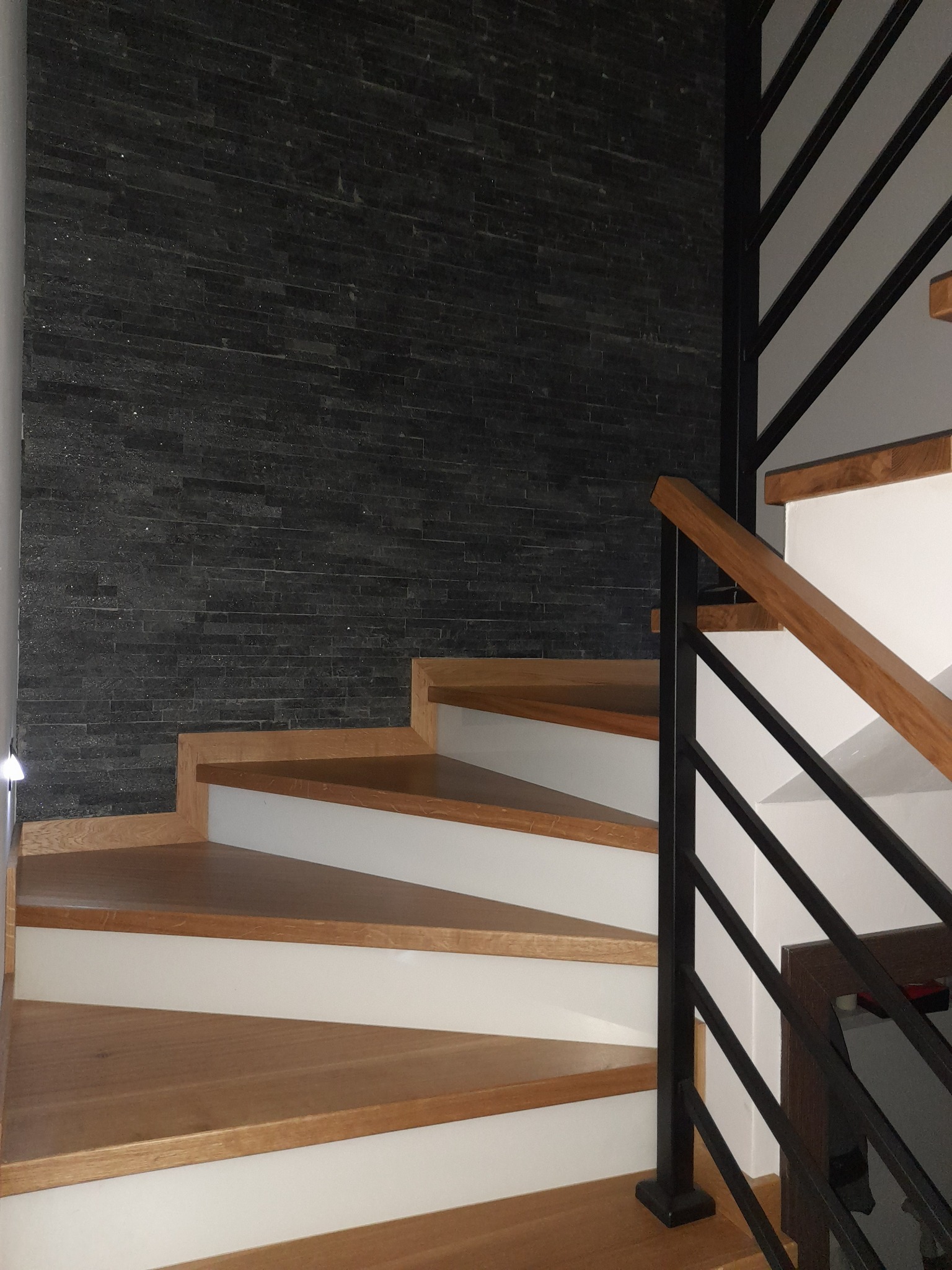 Schody drewniane z białymi podstopnicami i czarną metalową balustradą na tle ściany wykończonej czarnym kamieniem dekoracyjnym.