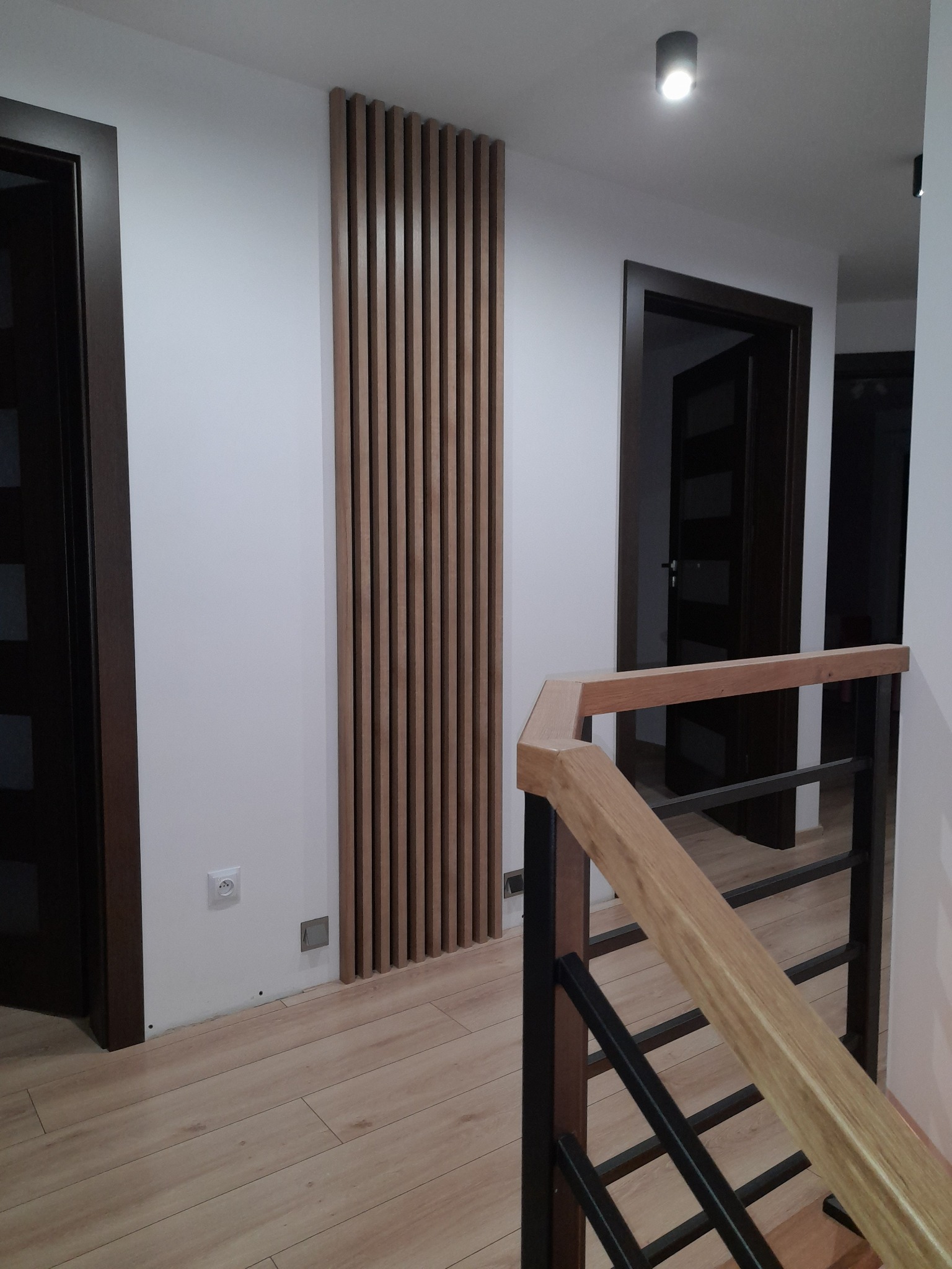 Nowoczesny przedpokój z drewnianą ażurową ścianką działową i schodami z drewnianą balustradą, widoczne drzwi wewnętrzne z ciemnego drewna.