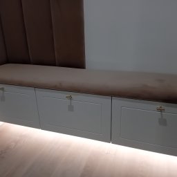 Usługi stolarskie Lazar Piotr - Siedzisko z szufladami w zabudowie, podświetlone od dołu, z brązowym obiciem i złotymi uchwytami. Minimalistyczny design, drewniana podłoga.