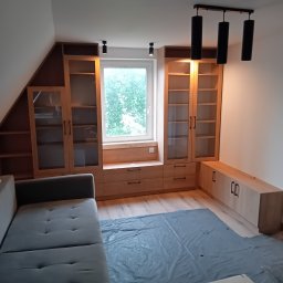 Usługi stolarskie Lazar Piotr - Jasna zabudowa meblowa z drewna w pokoju na poddaszu, z szafkami, półkami i szufladami przy oknie. Na podłodze folia ochronna. Nowoczesne oświetlenie sufitowe.