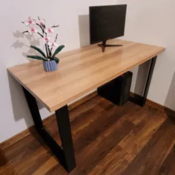 Drewniany blat biurka z czarnymi nogami w kształcie litery V, ustawiony na drewnianej podłodze, z monitorem i kwiatem w doniczce. Minimalistyczny design.