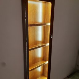 Usługi stolarskie Lazar Piotr - Wnęka ścienna z drewnianą, podświetloną półką, widoczna z boku. Ciemna rama i cztery półki z ciepłym oświetleniem tworzą unikalny element dekoracyjny.