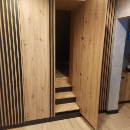 Usługi stolarskie Lazar Piotr - Drewniana szafa wnękowa z otwartymi drzwiami, ukazująca półki i schodki wewnątrz. Jasne drewno z czarnymi akcentami i pionowymi listwami. Widoczna podłoga i fragment ściany.