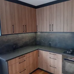 Nowoczesna kuchnia z drewnianymi frontami, czarnym blatem i sprzętem AGD. Uwagę zwracają minimalistyczne, podłużne uchwyty i ciemna listwa podsufitowa.