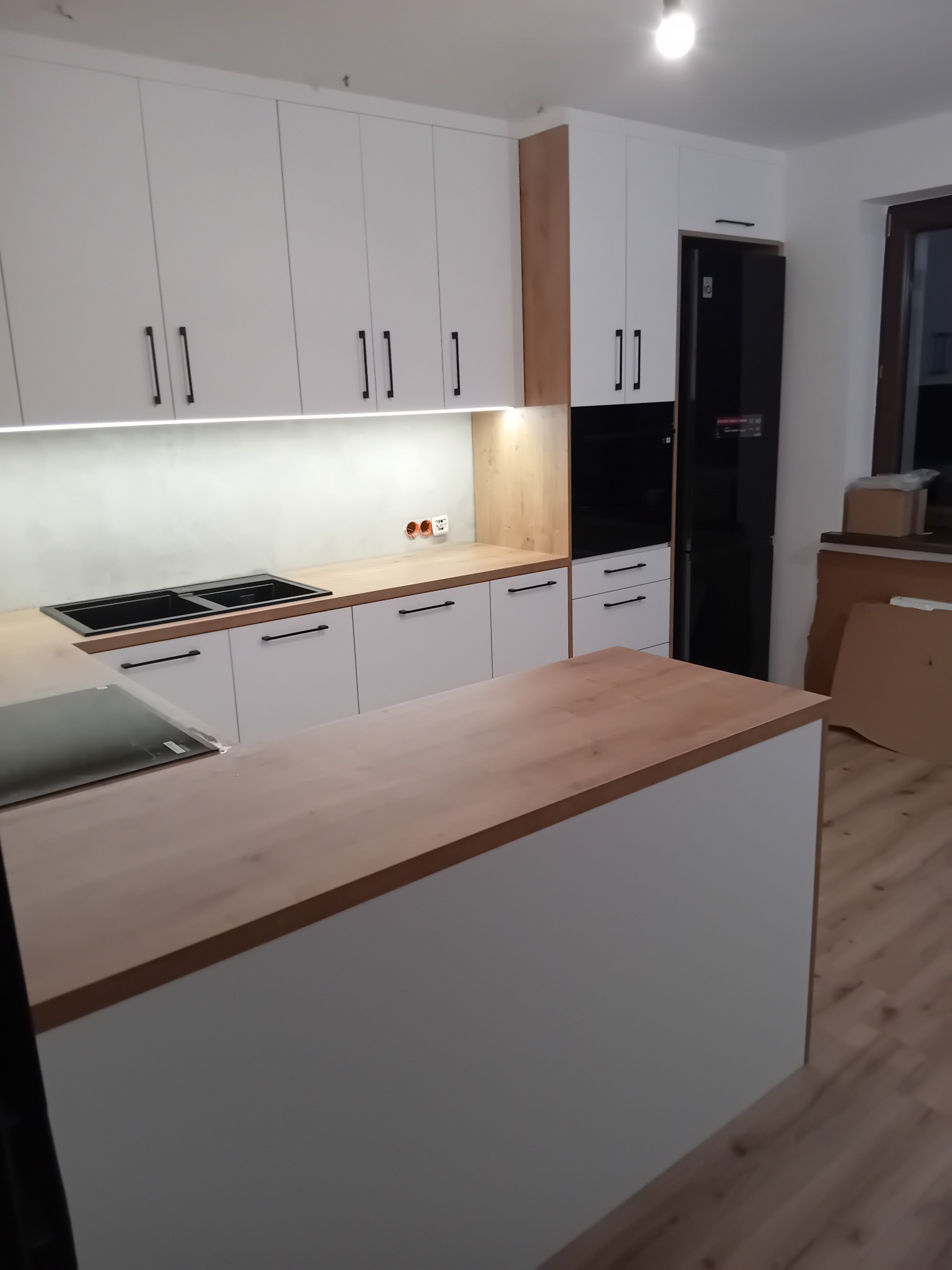 Nowoczesna kuchnia z białymi szafkami, drewnianym blatem i wyspą. Minimalistyczny design, czarne uchwyty i sprzęt AGD. Widoczne płyty indukcyjne i oświetlenie LED.