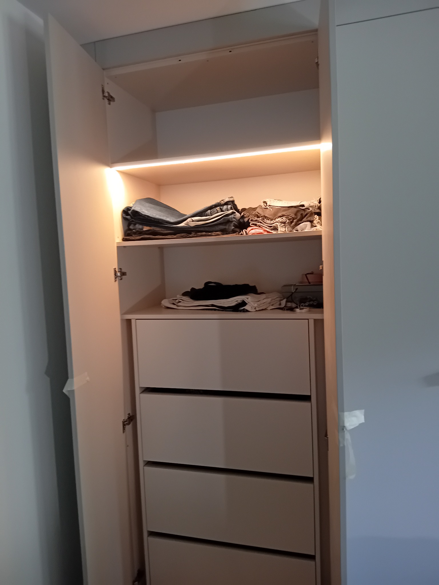 Otwarta szafa wnękowa z półkami i szufladami, oświetlona listwą LED. Widoczne ubrania na półkach. Fronty szuflad w kolorze kremowym, minimalistyczny design.