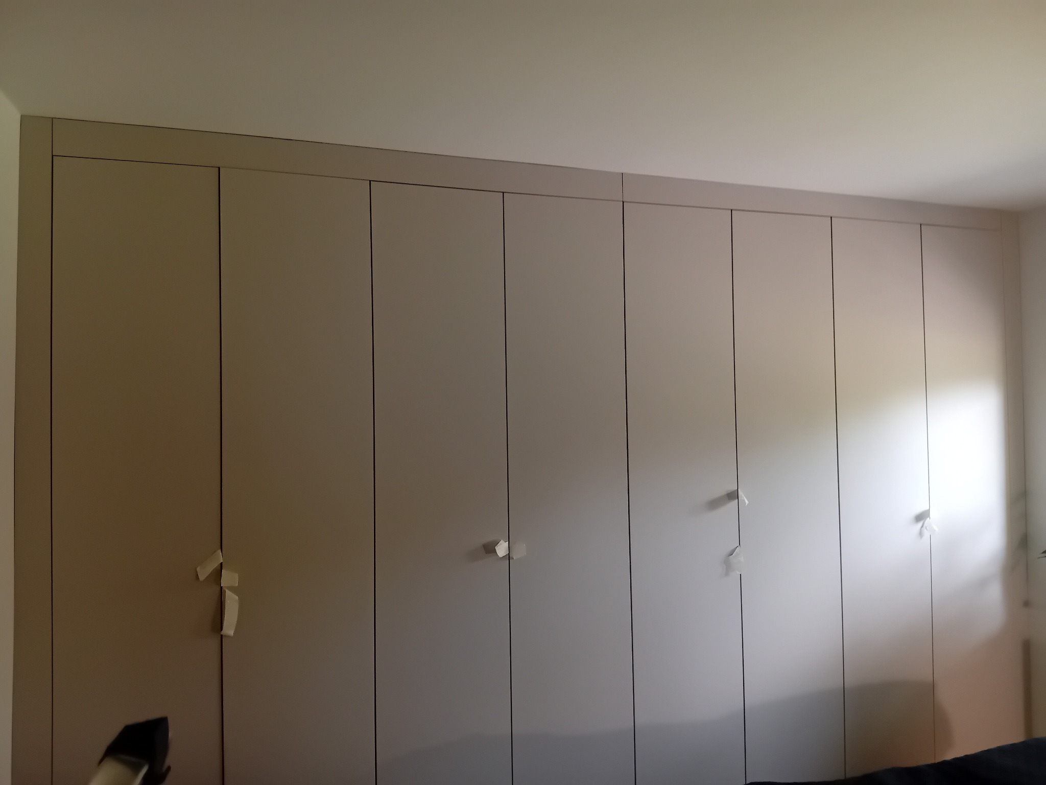 Beżowa szafa wnękowa z siedmioma drzwiami, z minimalistycznymi uchwytami zabezpieczonymi taśmą malarską. Widok na całą zabudowę od podłogi po sufit.