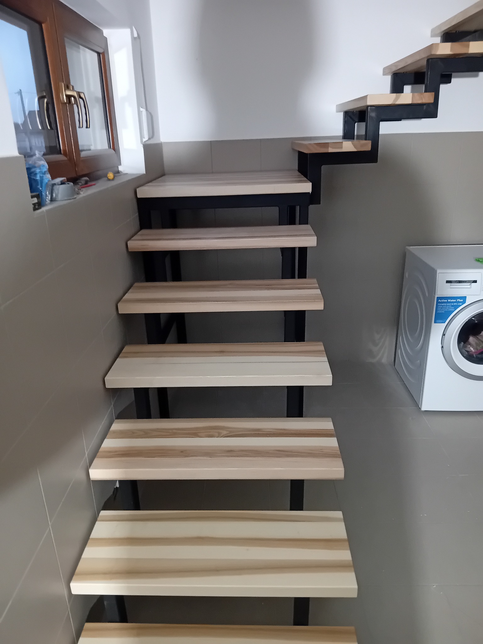 Nowoczesne schody drewniane z czarną, metalową konstrukcją, umieszczone w pomieszczeniu z pralką. Jasne drewno stopni kontrastuje z ciemnym metalem.