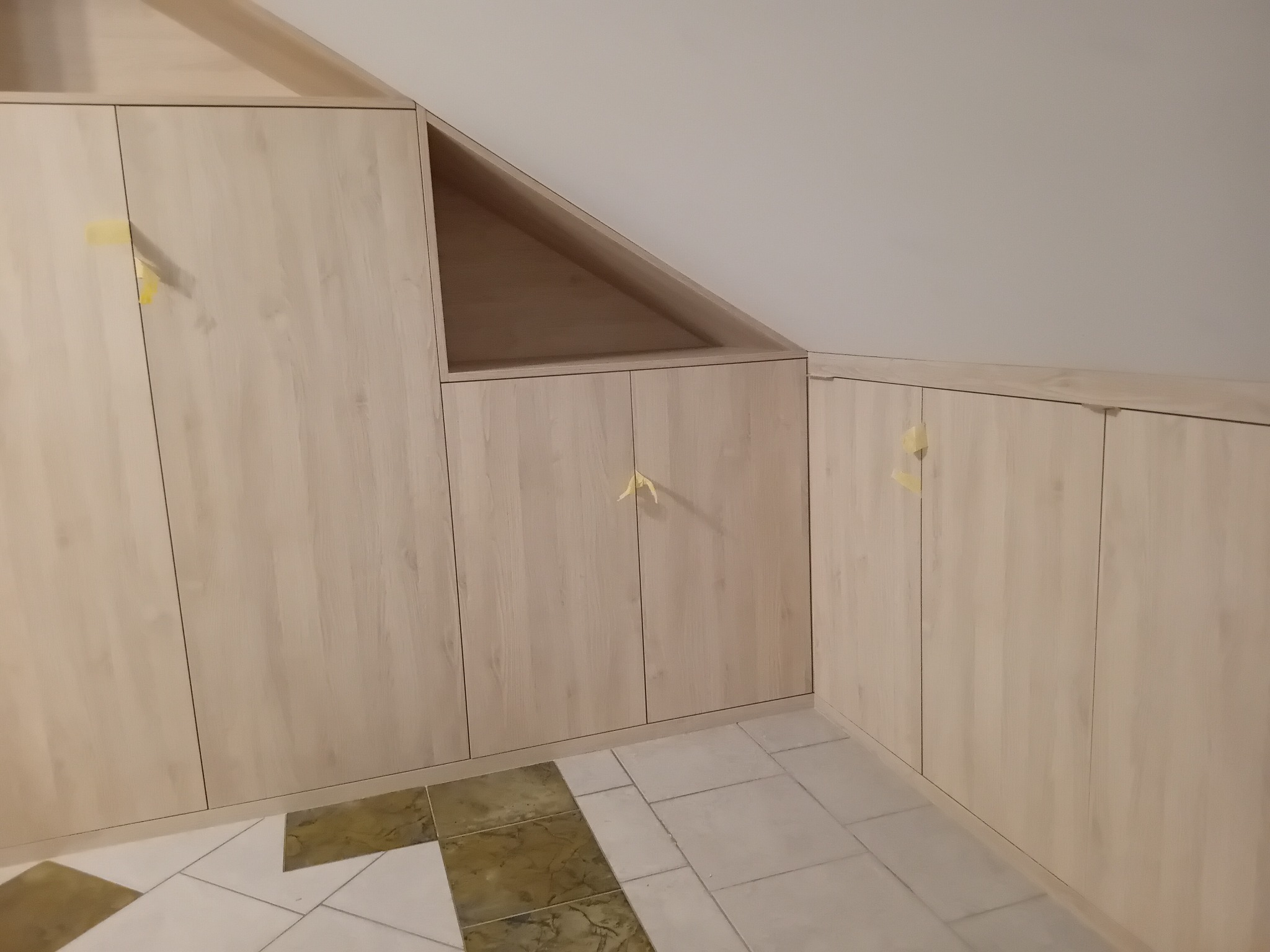Jasne, drewnopodobne szafy wnękowe zabudowane pod skosem dachu, z widocznymi żółtymi zabezpieczeniami na frontach, na tle beżowych płytek podłogowych.