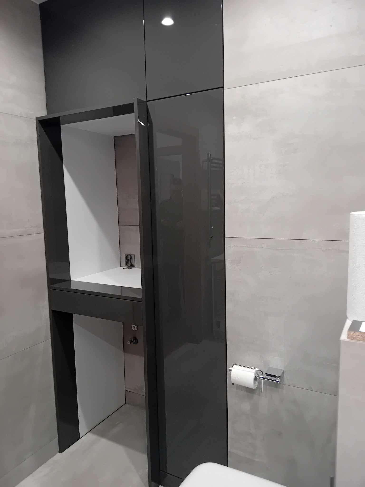 Nowoczesna szafka łazienkowa w odcieniach szarości z otwartą półką i błyszczącymi frontami, dopasowana do płytek na ścianie. Minimalistyczny design.