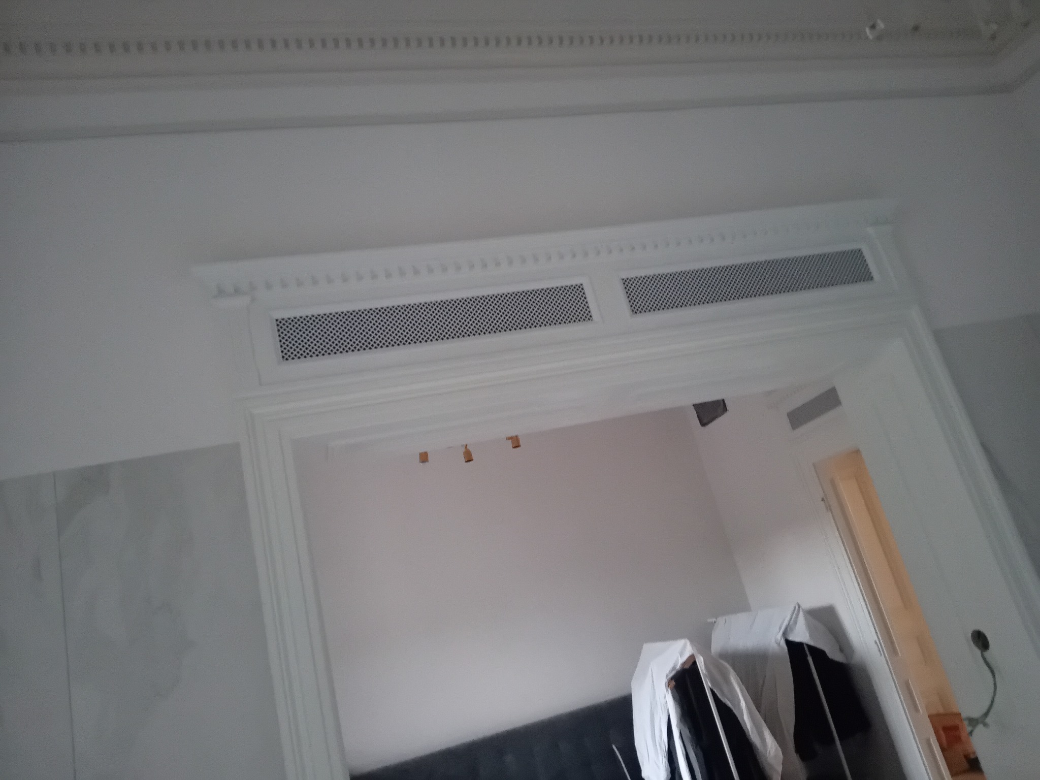 Białe, ozdobne obramowanie drzwi z ażurową kratką wentylacyjną nad przejściem. Widoczny fragment sufitu z dekoracyjną listwą i wnętrze pokoju.