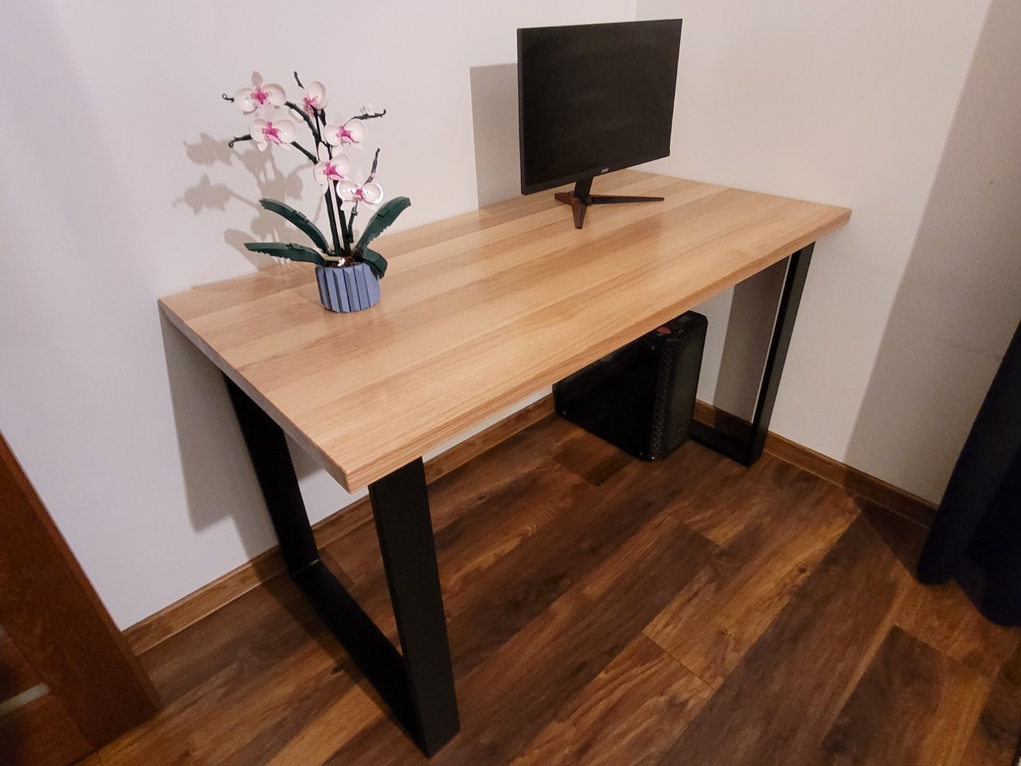 Drewniany blat biurka z czarnymi nogami w kształcie litery V, ustawiony na drewnianej podłodze, z monitorem i kwiatem w doniczce. Minimalistyczny design.