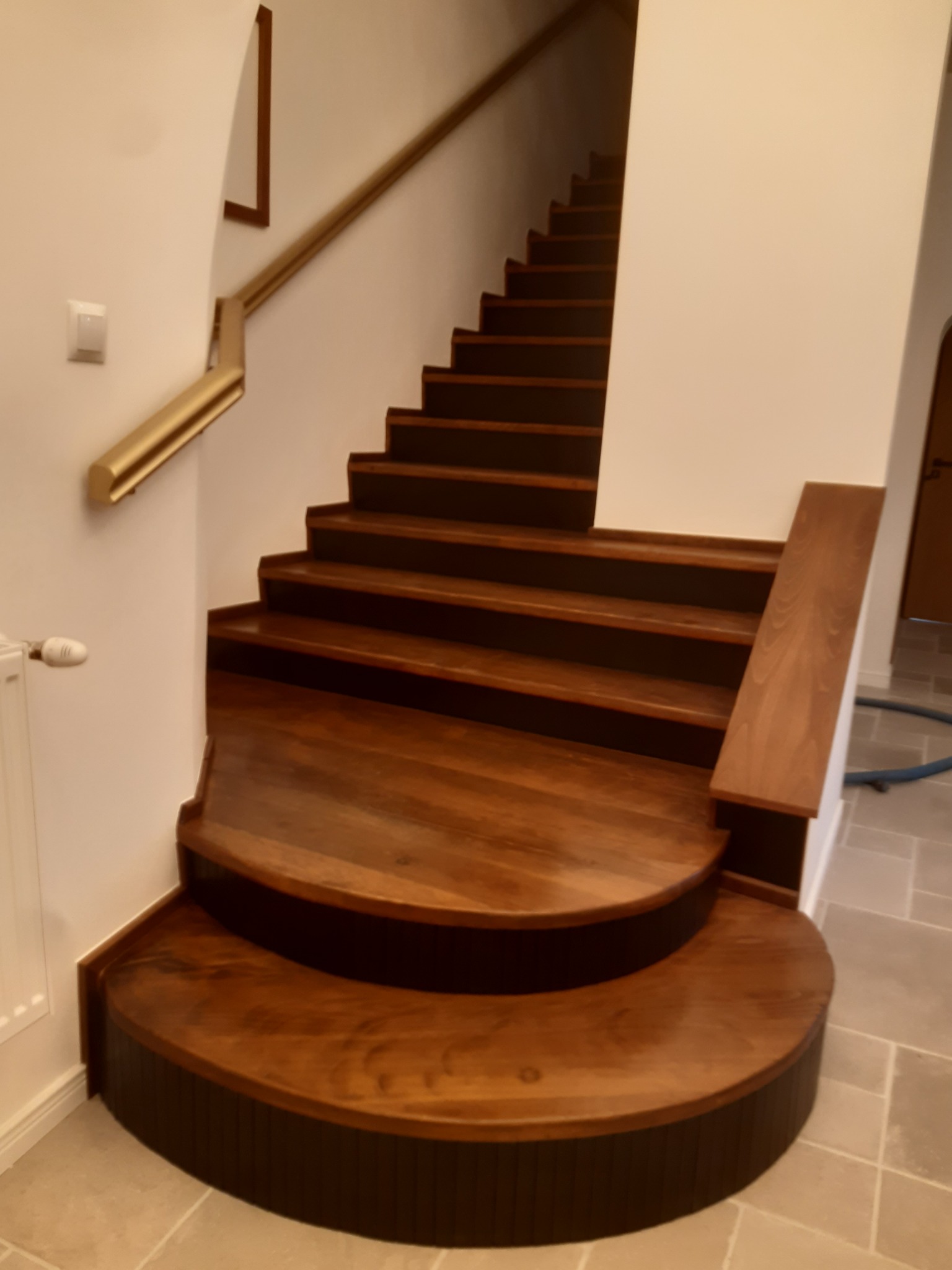 Drewniane schody z zaokrąglonymi stopniami i złotą balustradą, prowadzące w górę. Widoczne detale wykończenia i naturalne usłojenie drewna.