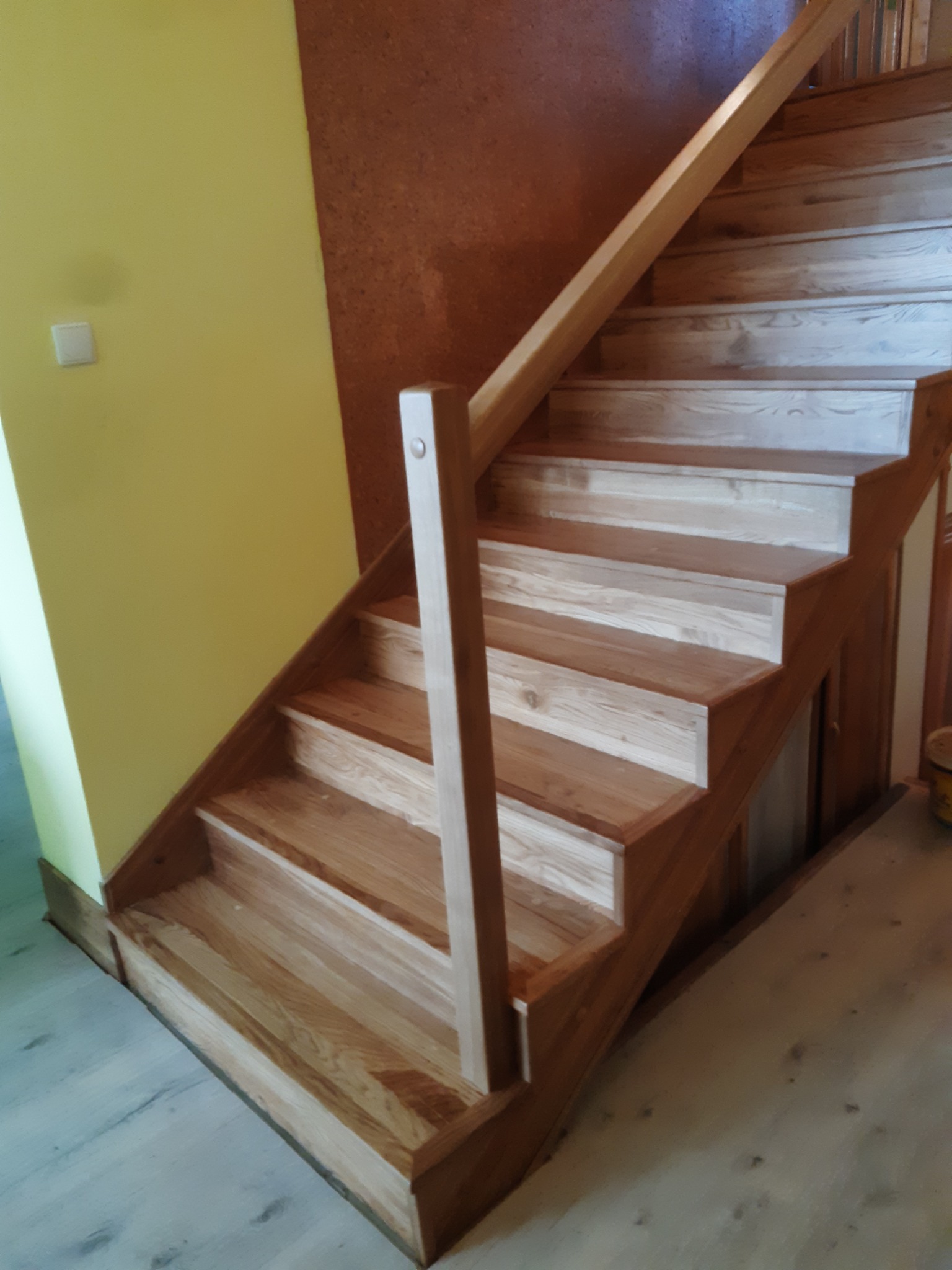 Jasne, drewniane schody z poręczą, umieszczone w narożniku pokoju z żółtą i brązową ścianą. Widoczne detale obróbki drewna i naturalny odcień materiału.