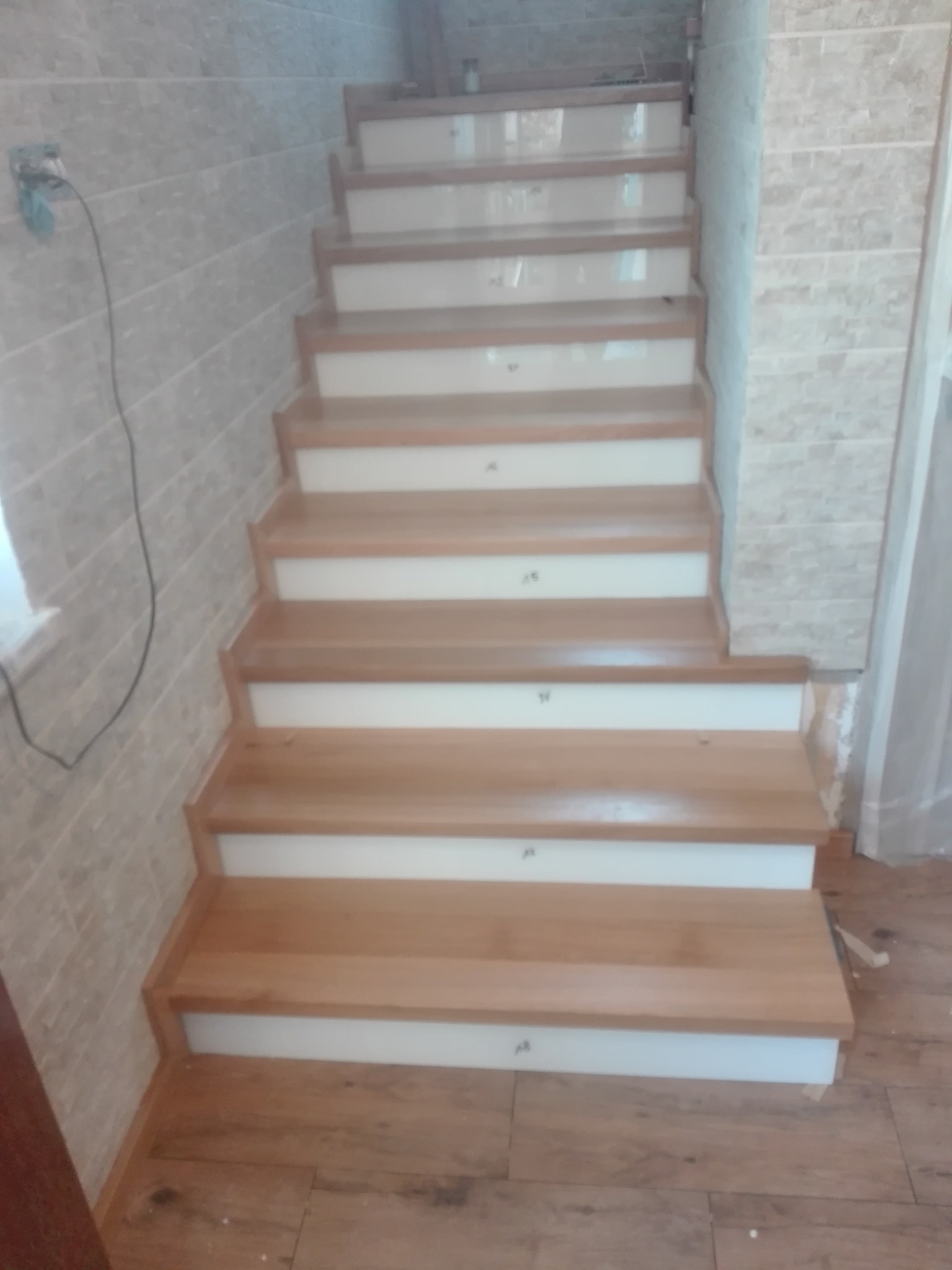 Jasne drewniane schody z białymi podstopnicami, widok z dołu. Ułożone schody wewnątrz budynku, ściany wyłożone płytkami imitującymi cegłę.