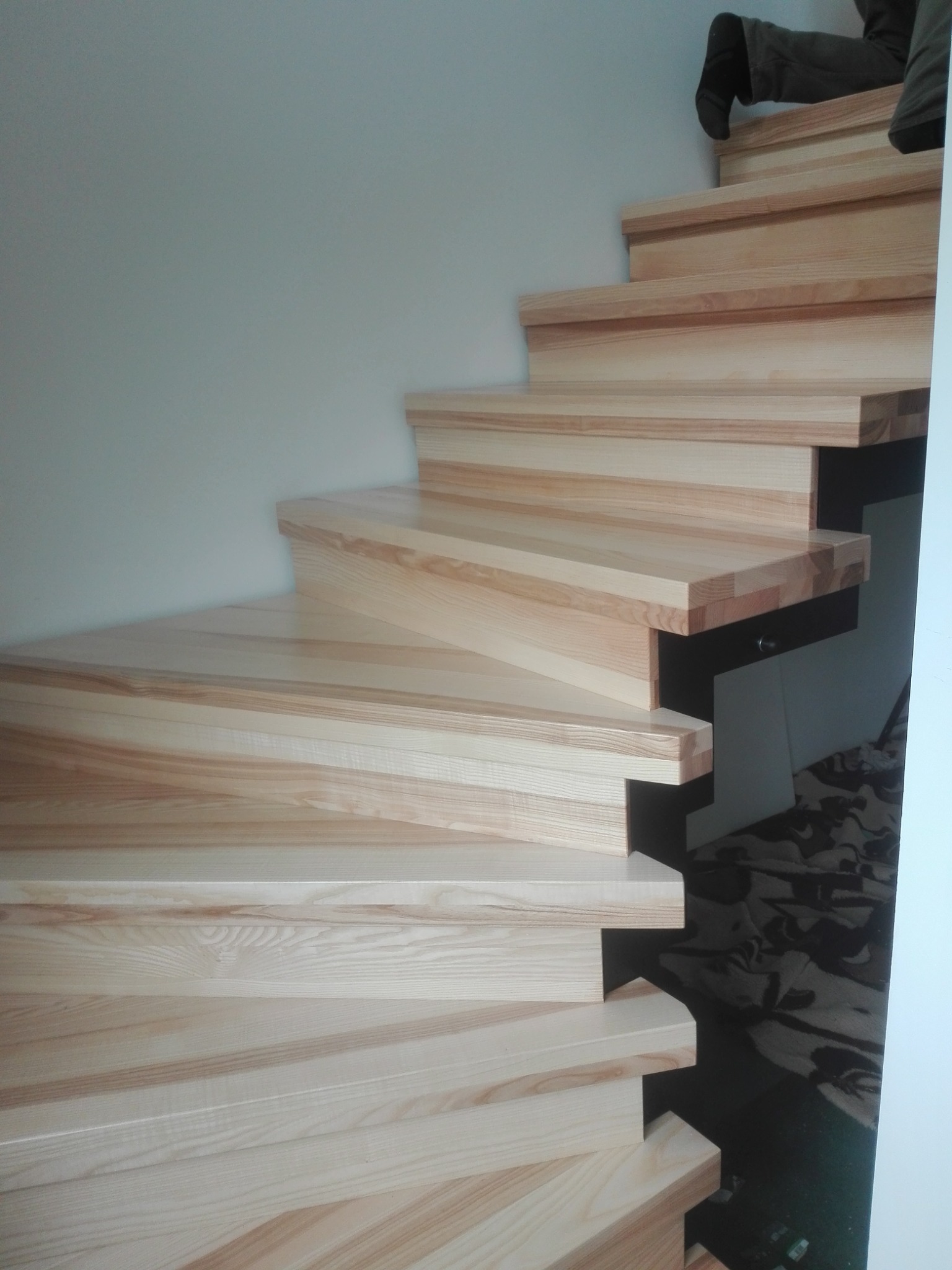 Jasne, drewniane schody z widocznym usłojeniem drewna, prowadzące w górę. Na górnych stopniach widoczne fragmenty osoby. Minimalistyczny design.