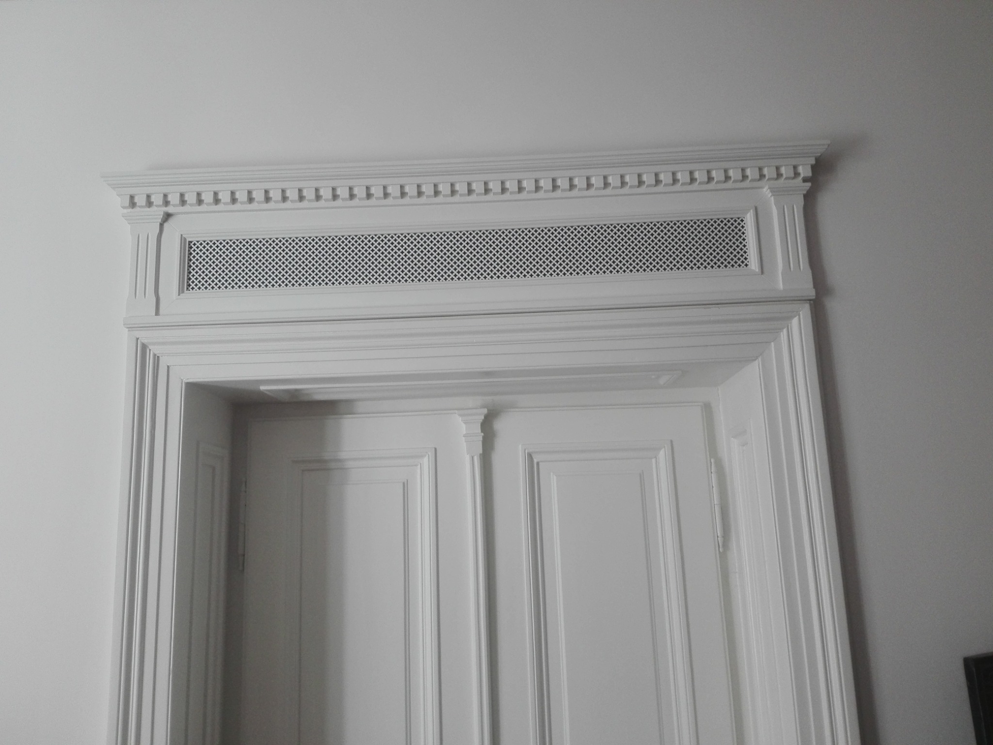 Białe, klasyczne drzwi z dekoracyjną ażurową listwą wentylacyjną nadproża. Dwoje drzwi panelowych z ozdobnymi listwami i pilastrami.