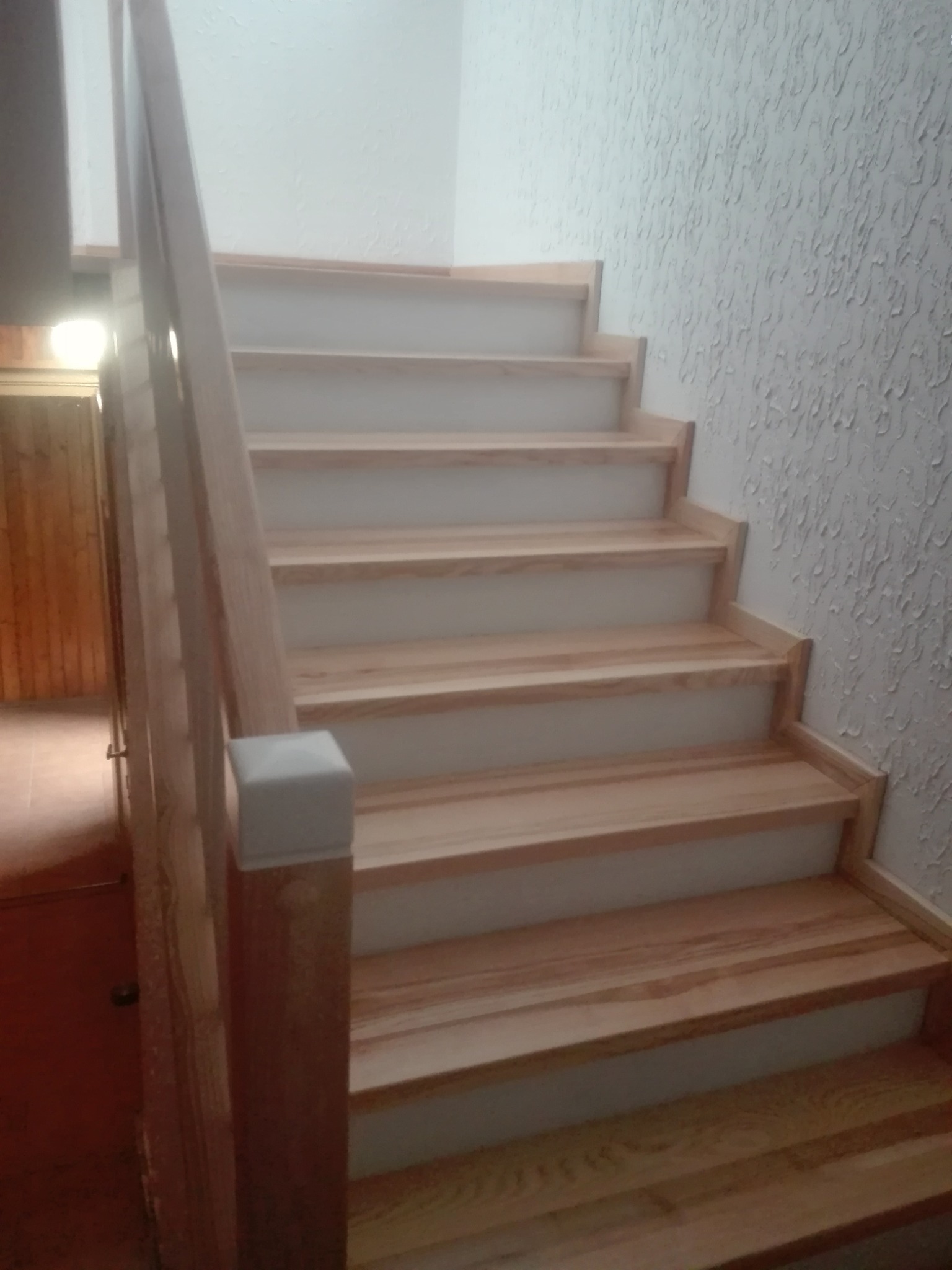 Jasne drewniane schody z białymi podstopnicami i drewnianą balustradą, widoczne z dołu, na tle ściany o fakturze baranka.