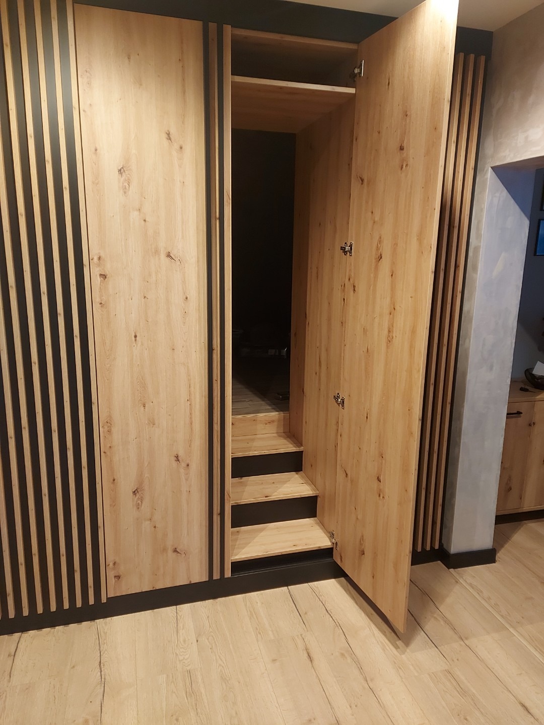 Drewniana szafa wnękowa z otwartymi drzwiami, ukazująca półki i schodki wewnątrz. Jasne drewno z czarnymi akcentami i pionowymi listwami. Widoczna podłoga i fragment ściany.