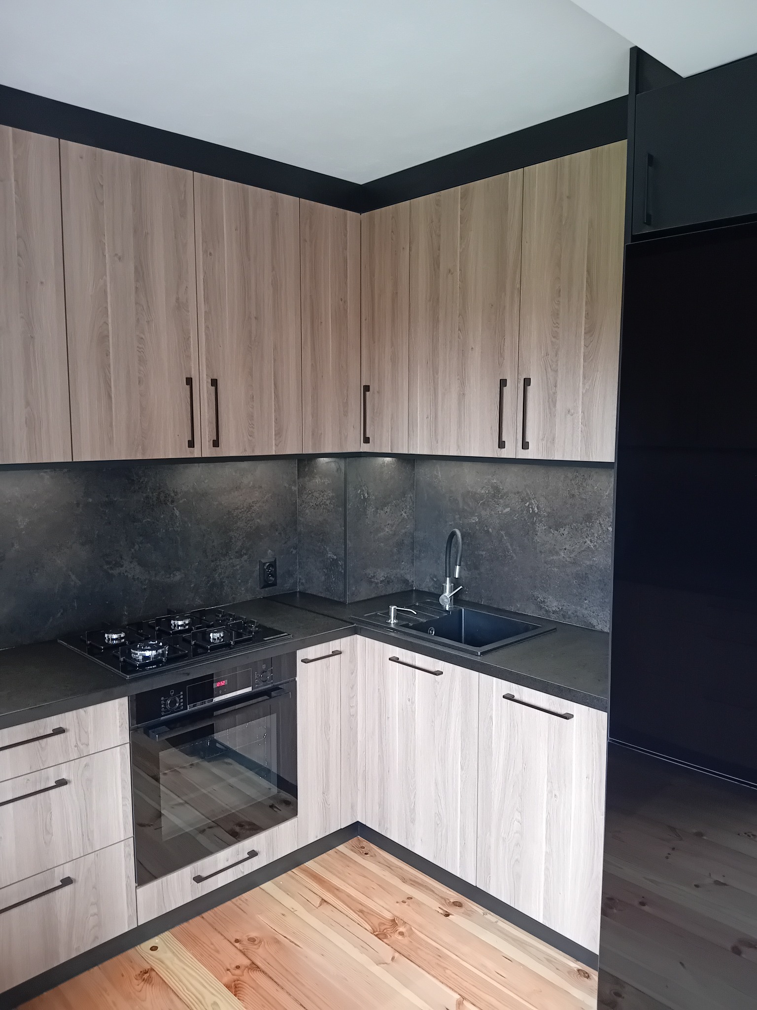 Nowoczesna kuchnia narożna z drewnianymi frontami i czarnymi akcentami: blaty, listwa podszafkowa, uchwyty. Widoczna płyta grzewcza i piekarnik.