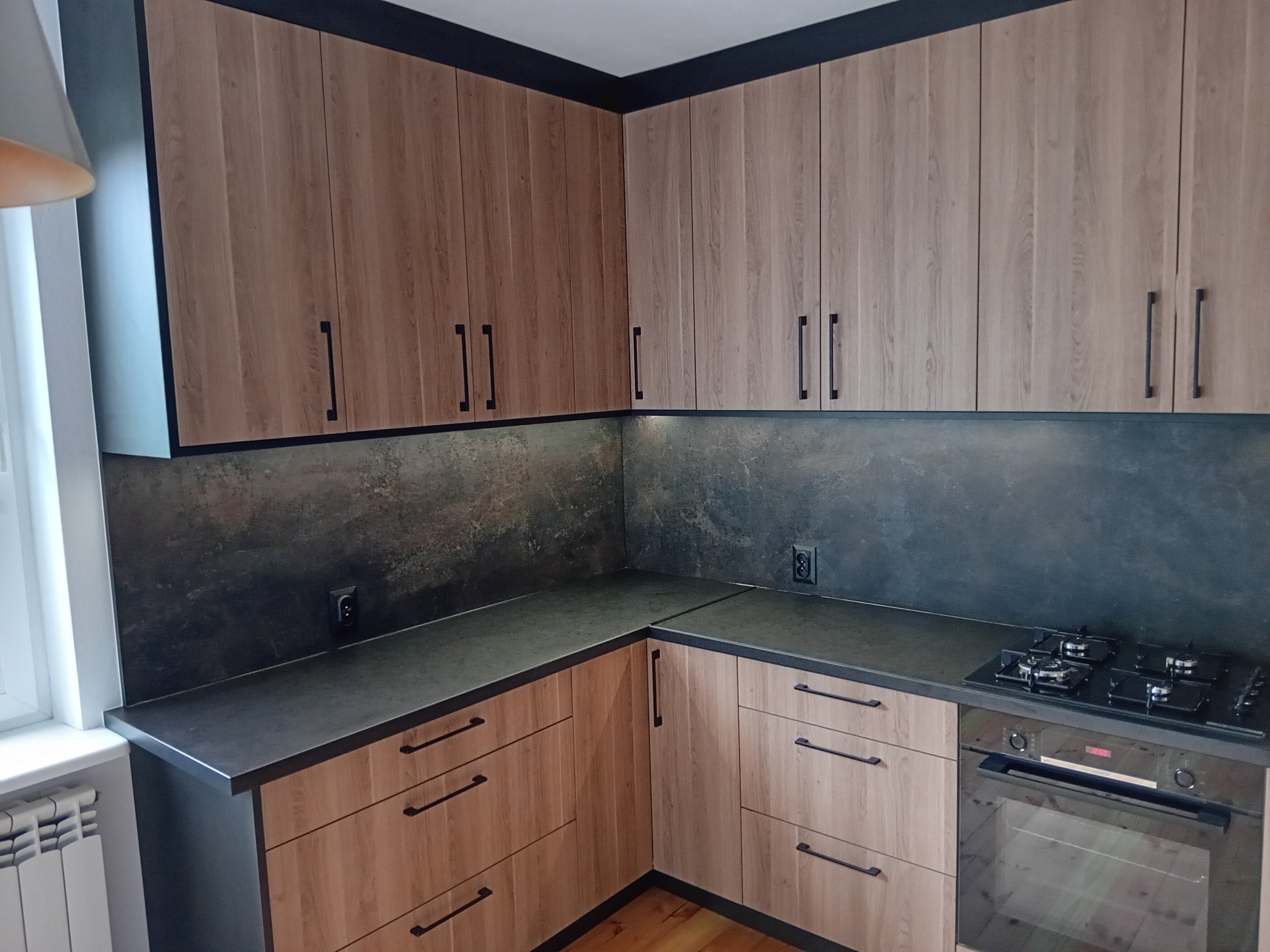 Nowoczesna kuchnia z drewnianymi frontami, czarnym blatem i sprzętem AGD. Uwagę zwracają minimalistyczne, podłużne uchwyty i ciemna listwa podsufitowa.