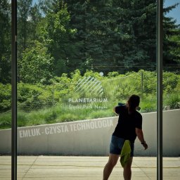 Klein - Osoba w trakcie czyszczenia dużej szklanej powierzchni z napisem 'PLANETARIUM Śląski Park Nauki' oraz 'EMLUK - CZYSTA TECHNOLOGIA', widok zza szyby na zieleń drzew.
