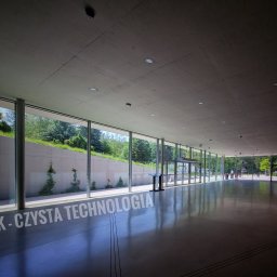 Klein - Wnętrze nowoczesnego budynku z przeszkloną ścianą, widok na zieleń na zewnątrz, na podłodze napis 'EMLUK - CZYSTA TECHNOLOGIA'.