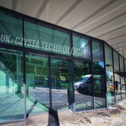 Klein - Mycie szklanej fasady budynku z napisem 'EMLUK - CZYSTA TECHNOLOGIA' za pomocą lancy teleskopowej i szczotki, widoczny samochód dostawczy i osoba w tle.