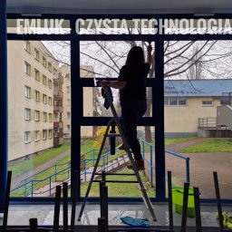 Klein - Kobieta na drabinie czyści okno z logo firmy 'EMLUK CZYSTA TECHNOLOGIA', widok z wnętrza pomieszczenia na budynki mieszkalne i drzewa.