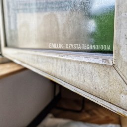 Klein - Bardzo brudne okno przed czyszczeniem, widoczne z bliska z naklejonym logo firmy 'EMLUK - CZYSTA TECHNOLOGIA'.
