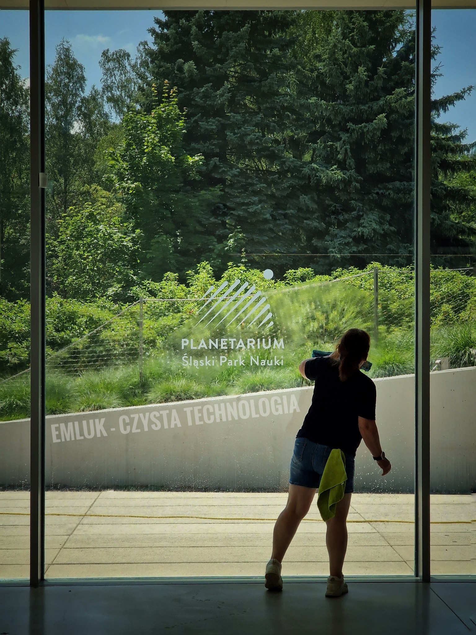 Osoba w trakcie czyszczenia dużej szklanej powierzchni z napisem 'PLANETARIUM Śląski Park Nauki' oraz 'EMLUK - CZYSTA TECHNOLOGIA', widok zza szyby na zieleń drzew.